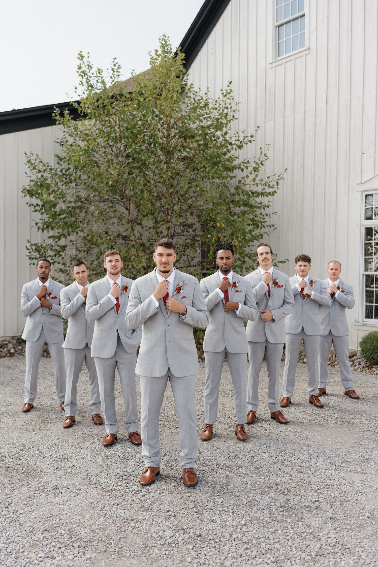 Groomsmen in light gray suits and brown shoes pose playfully in a line against a white barn wall.
