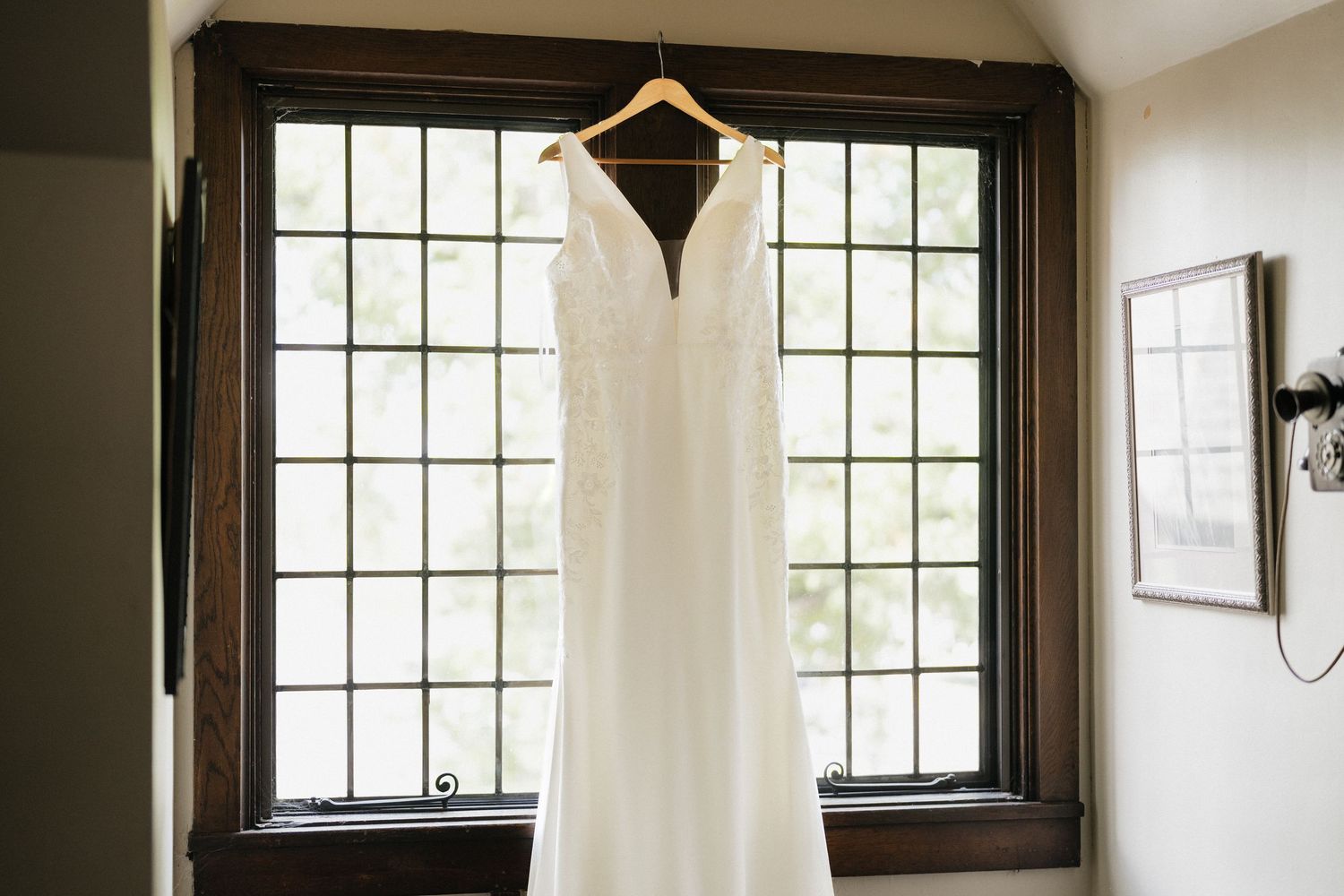 A white wedding dress hangs elegantly by a window with dark trim in natural light.