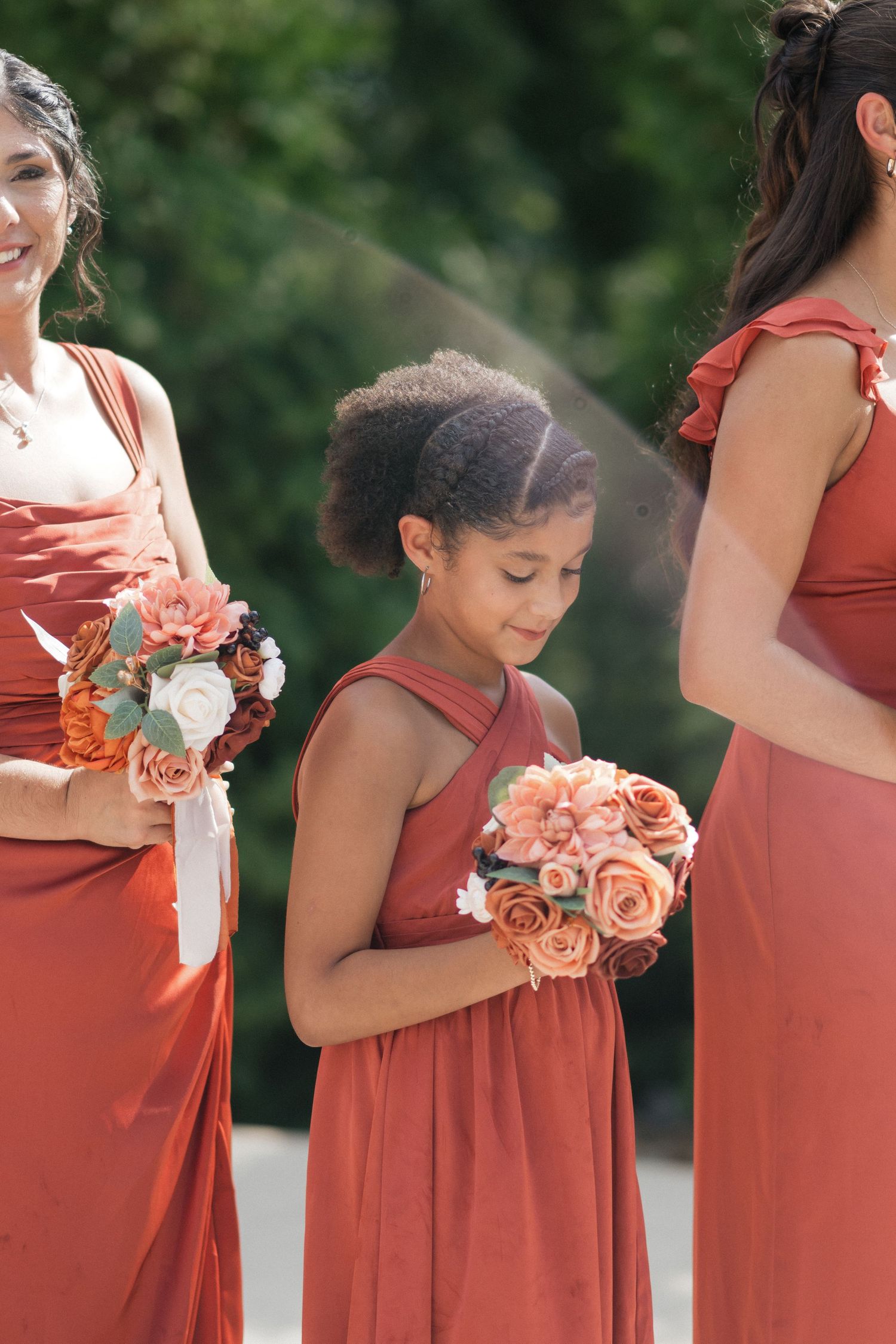 Bridesmaids in rust-colored dresses holding peach rose bouquets at outdoor wedding.