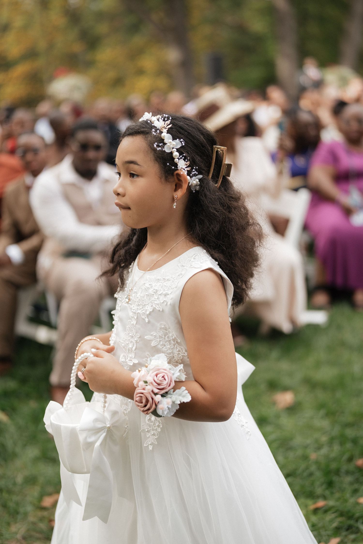 Young flower girls in white lace dresses with floral details walk together on grass.