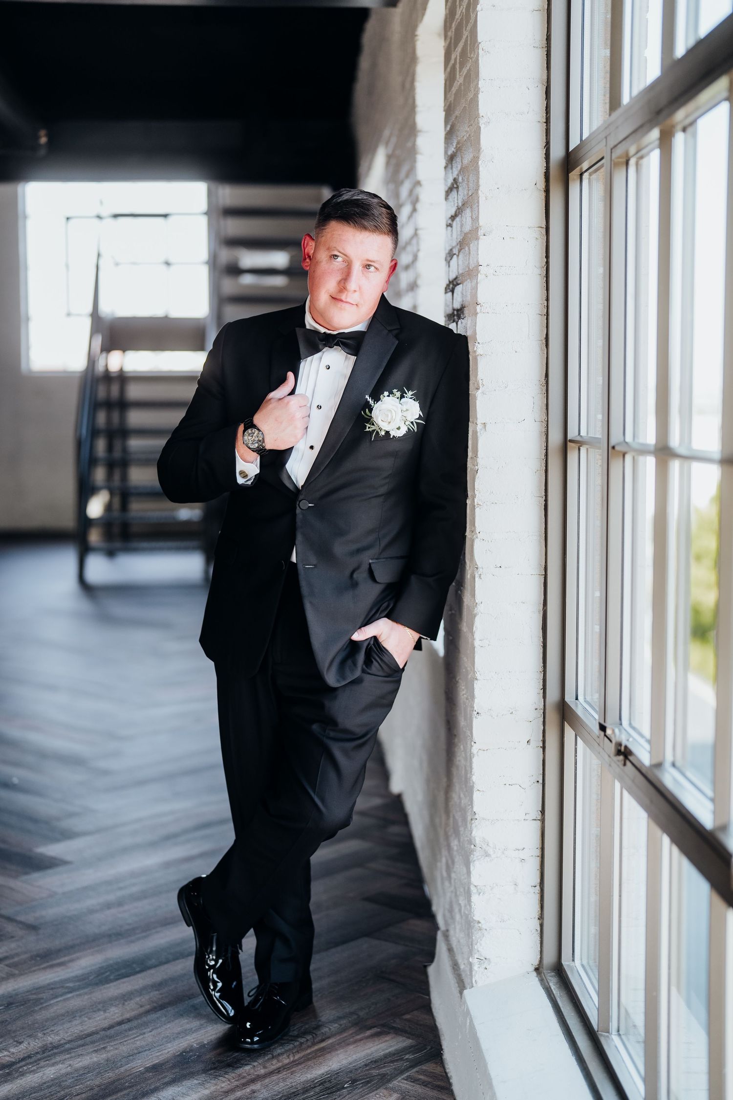 Groom in black tuxedo posing by large windows for formal portraits.