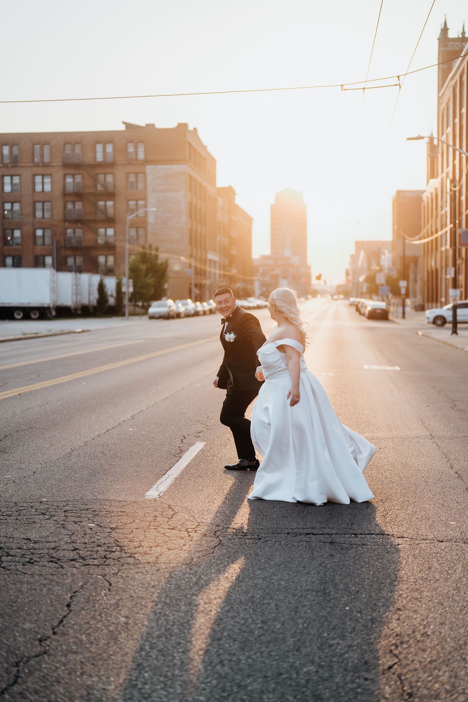 A romantic wedding couple dances in an urban street at sunset with golden light streaming between buildings.