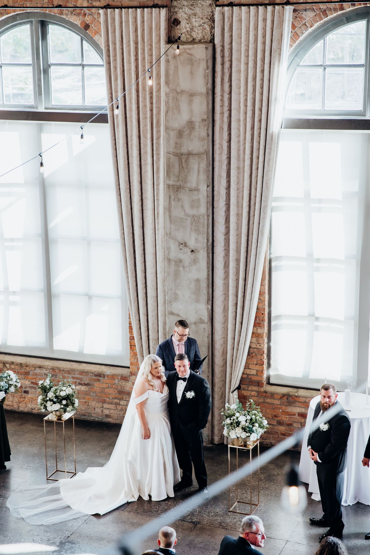 Wedding couple exchanges vows in a modern industrial venue with arched windows and brick walls.