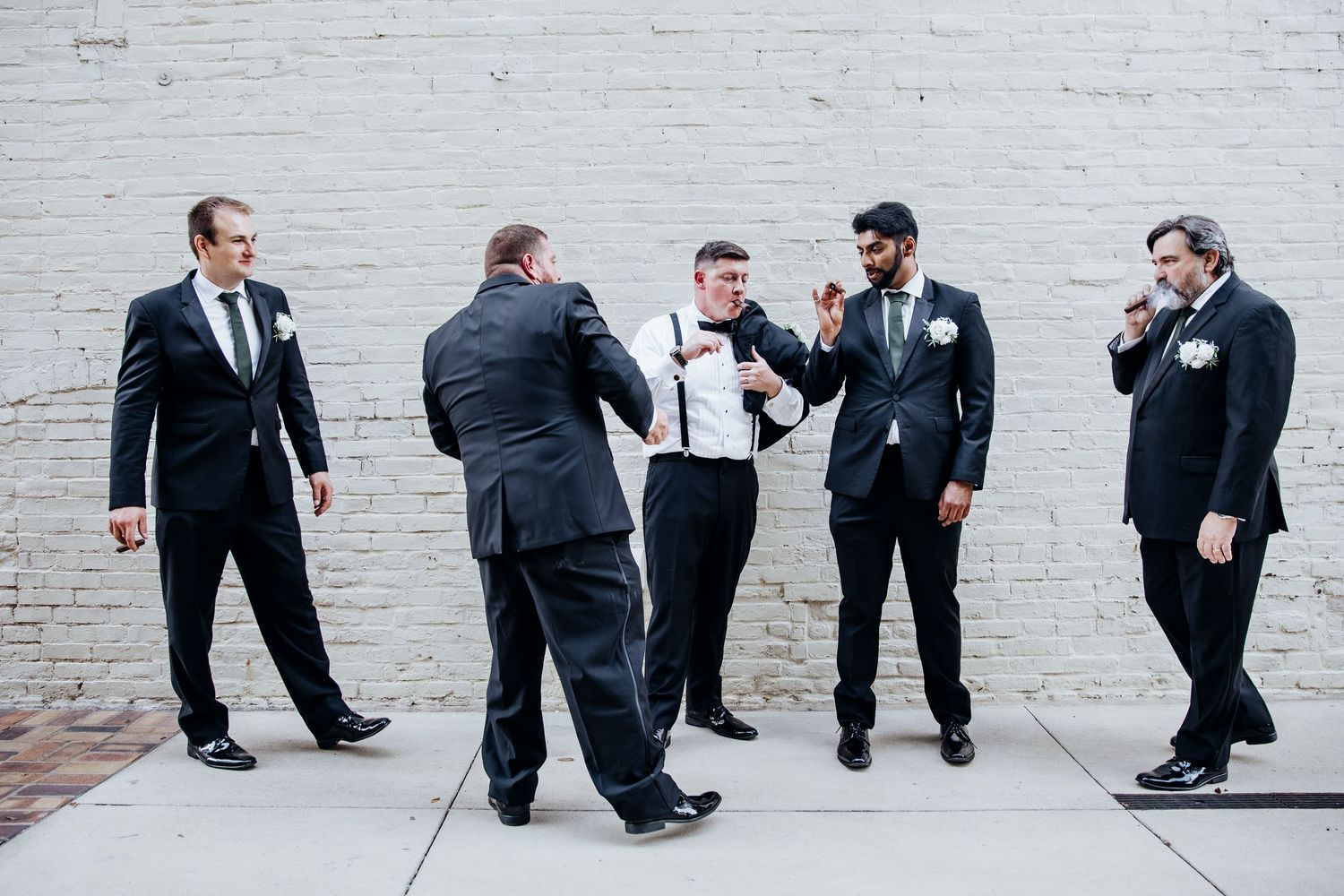 Wedding party members in formal attire posing casually against a white brick wall.