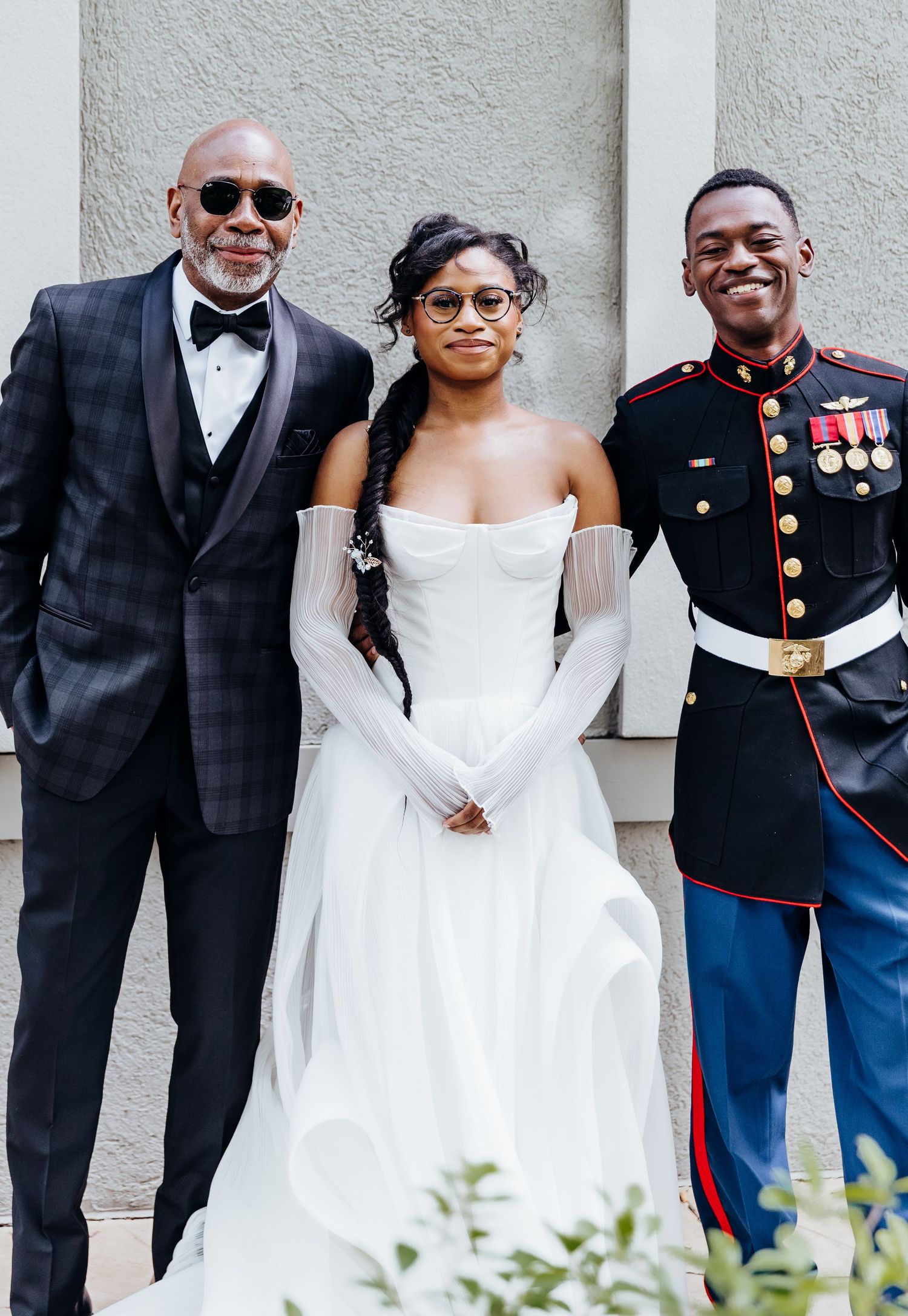 A wedding party poses together against a wall, featuring formal attire and a military uniform.