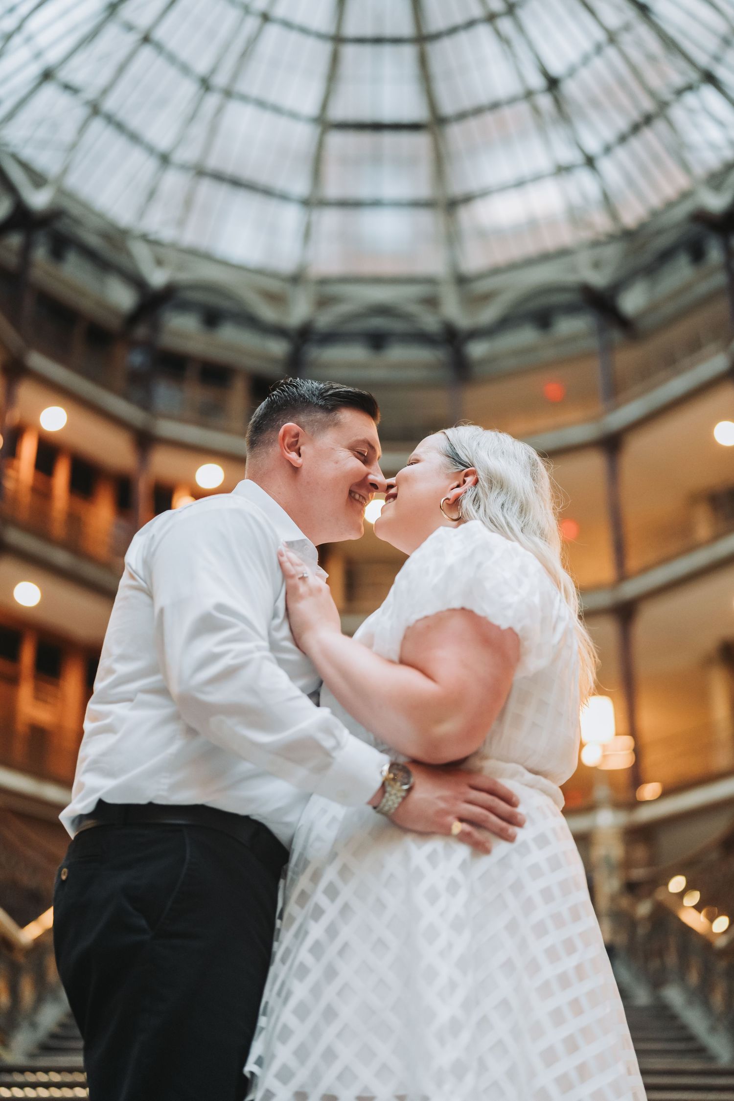 A romantic moment captured on an ornate staircase under a glass dome ceiling.