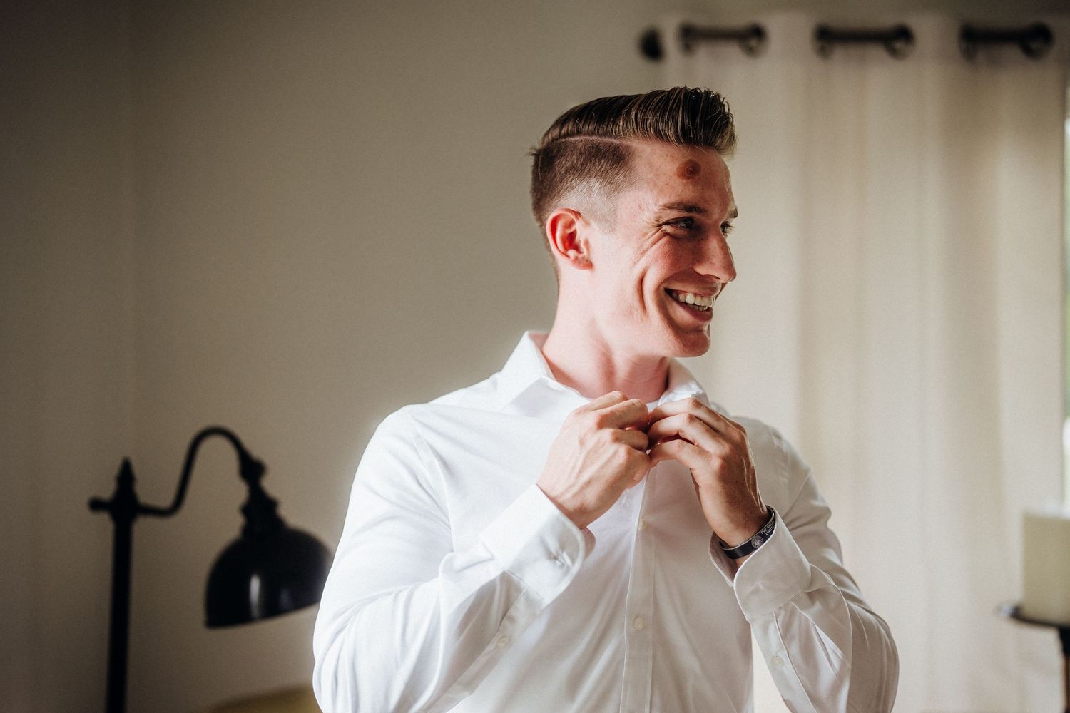 A groom in a white dress shirt adjusts his bow tie while getting ready.