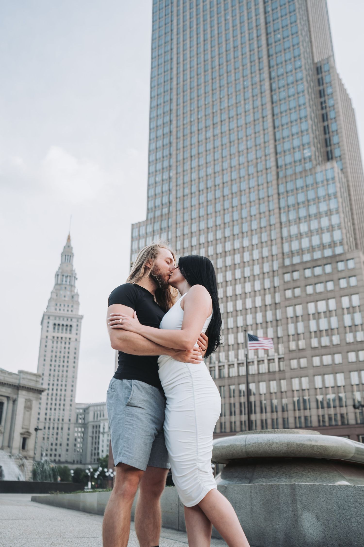 A couple embraces in front of a modern skyscraper and historic building in an urban setting.