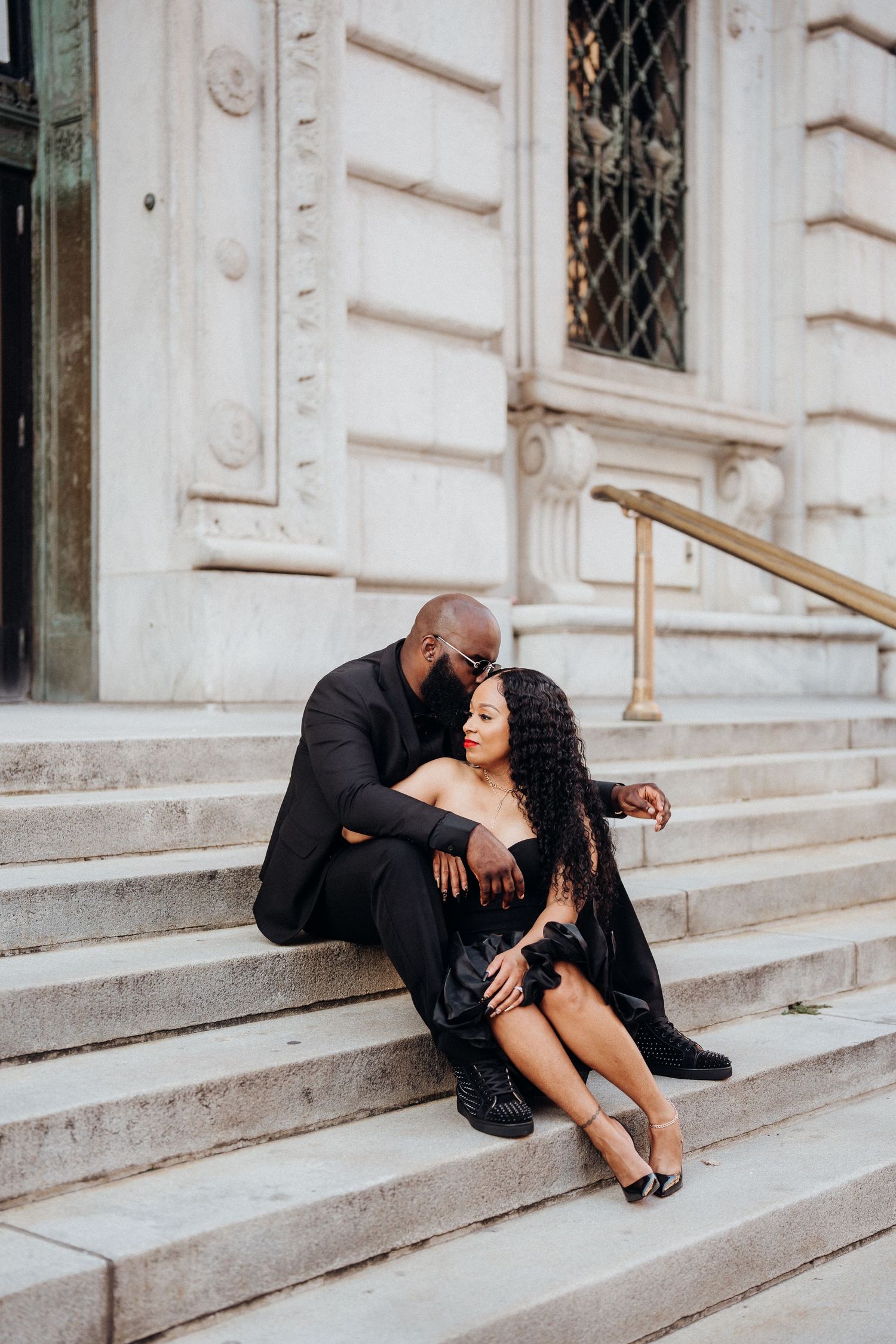 A couple embraces on elegant stone steps in formal black attire.