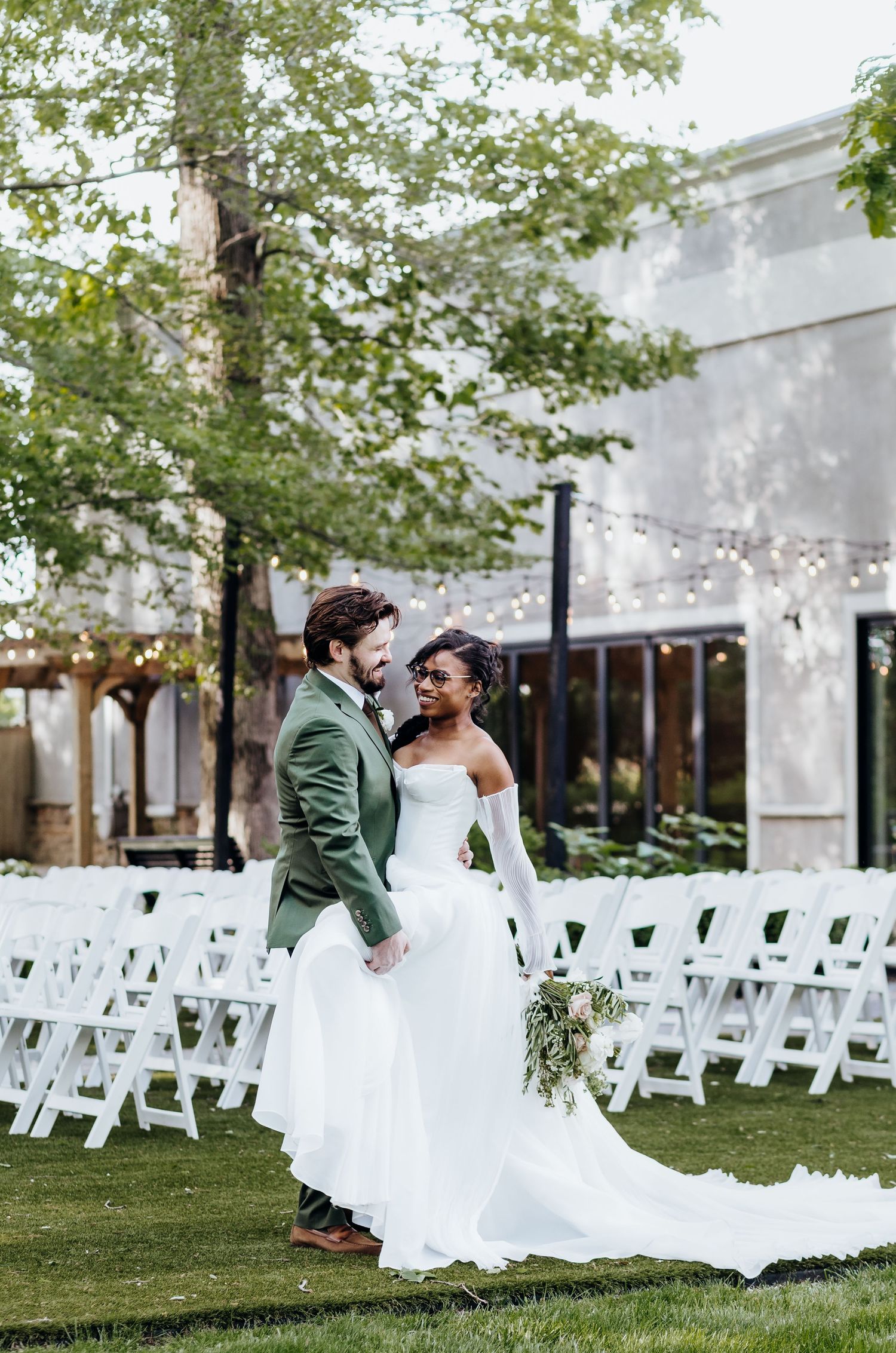 Couple shares romantic moment on lawn with white ceremony chairs and string lights in background.