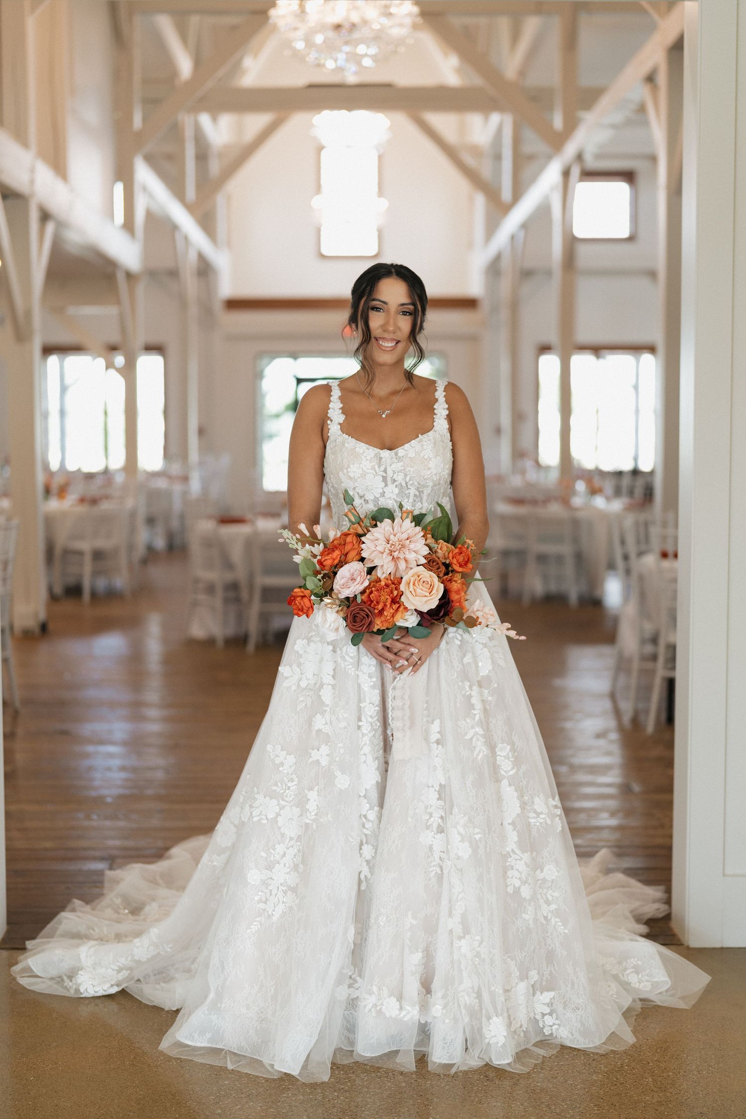 A bride in a white lace wedding gown stands in an elegant white indoor venue with tall columns.