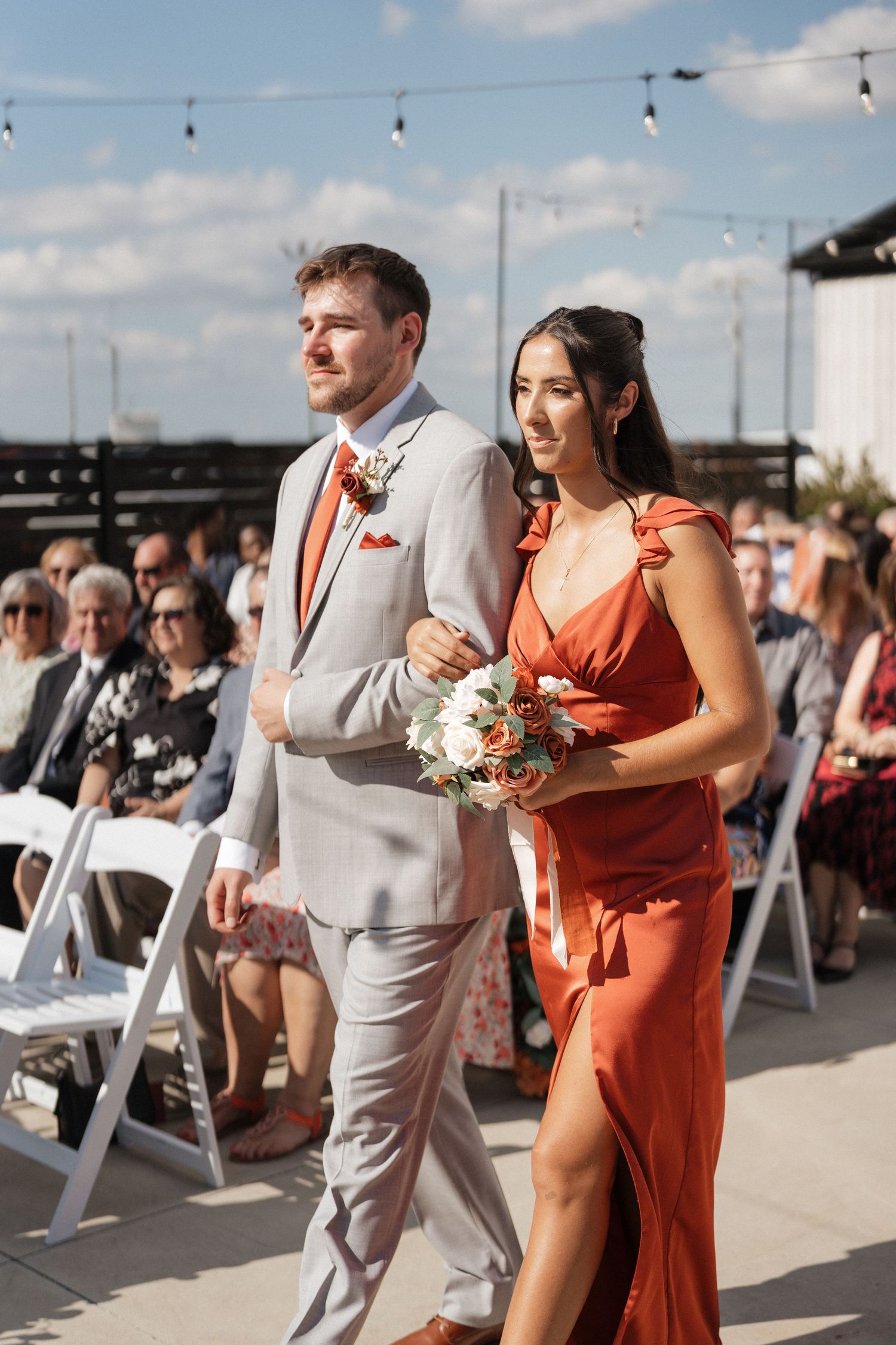 Wedding processional walking outdoors with grey suits and rust-colored dresses against blue sky.