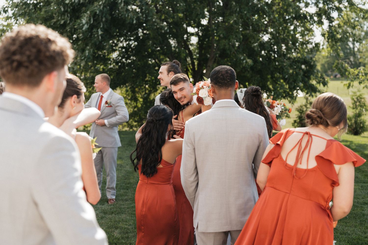 Wedding party in grey suits and rust-colored dresses poses outdoors on a sunny day.