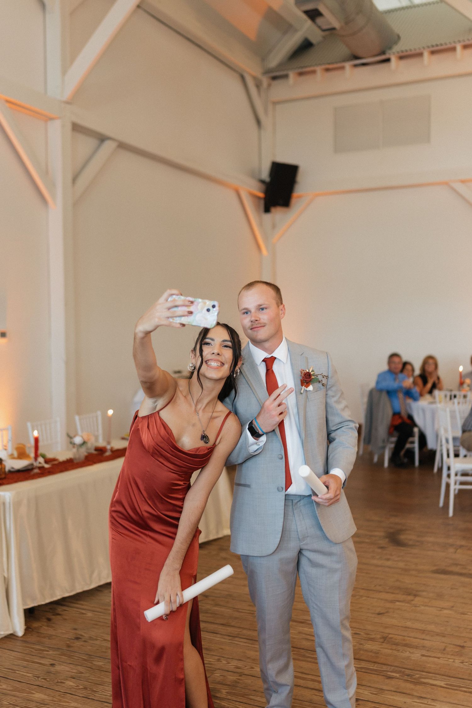 Wedding guests take selfie photos during reception in elegant white venue with wooden beams.