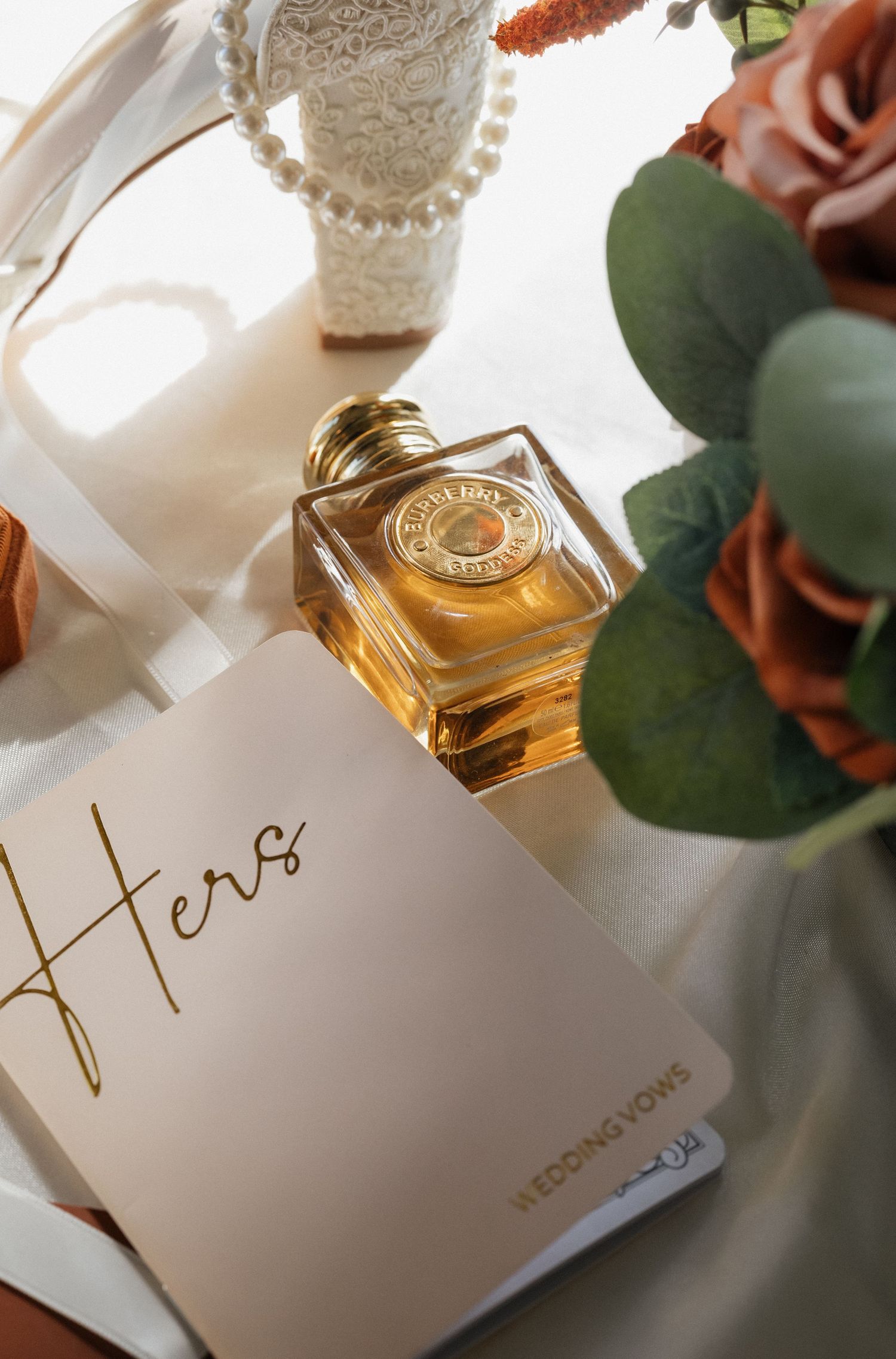 Wedding invitation details and perfume bottle arranged with eucalyptus leaves and floral elements on white surface.