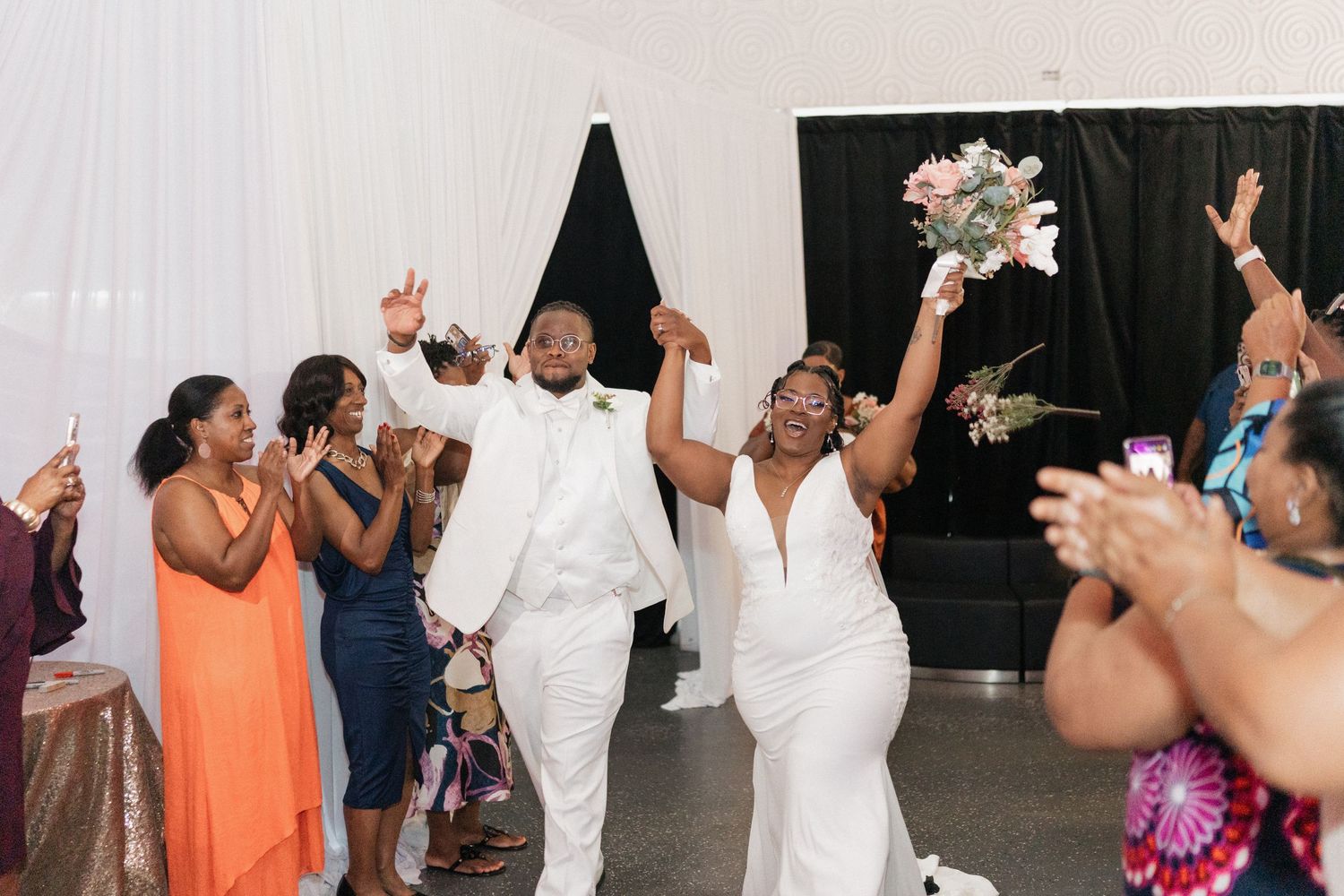 Newlyweds celebrate with guests during their wedding reception entrance with raised arms and cheers.