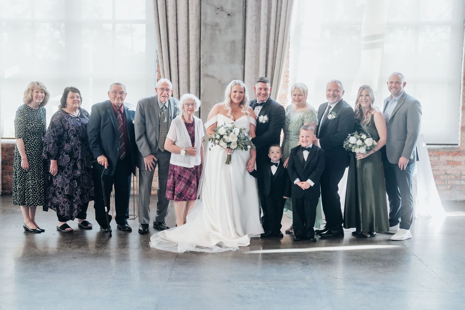 Wedding party and guests posed together in a formal group photo against a light-colored wall.