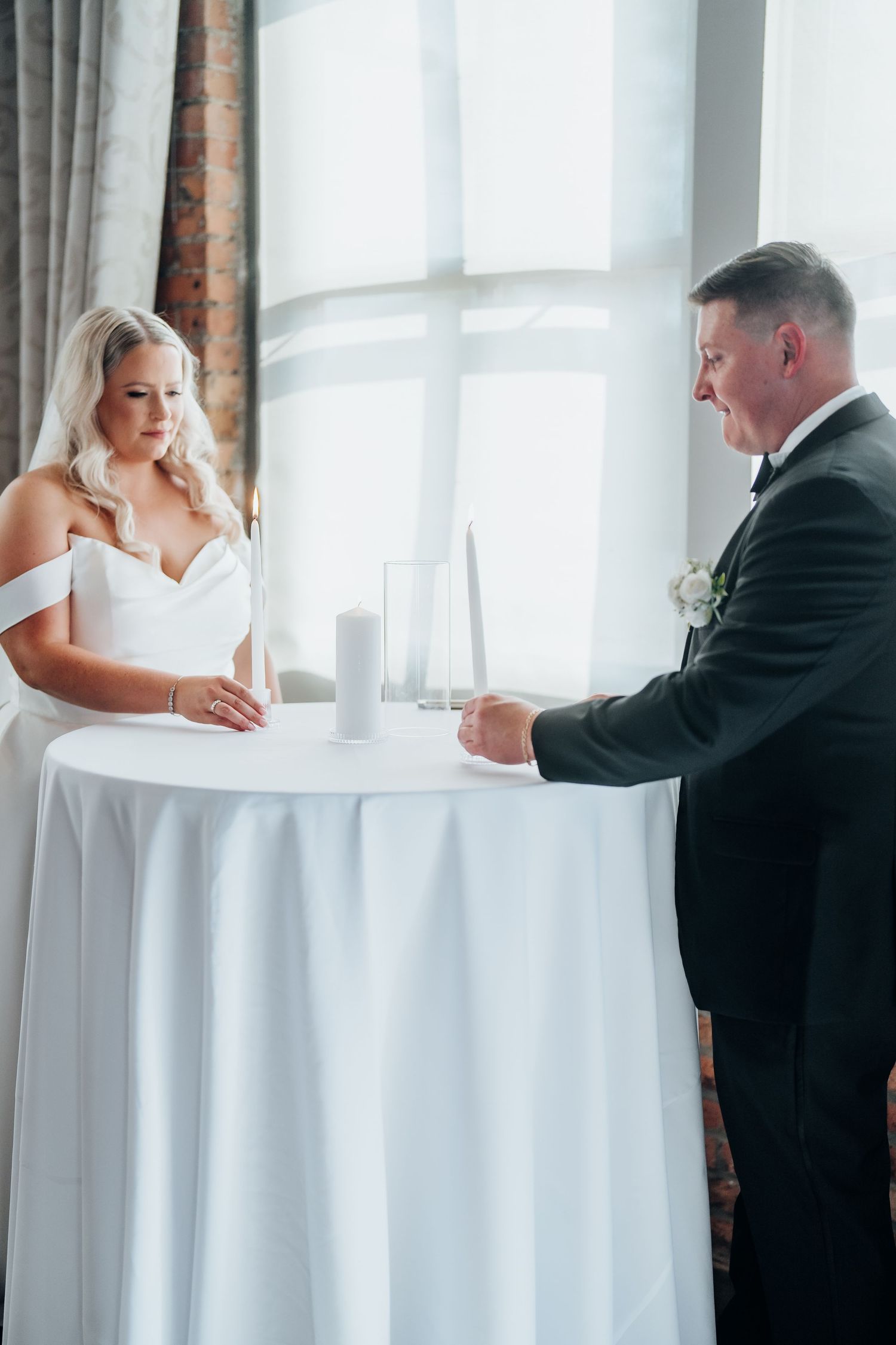 Wedding party members signing official documents at a white-clothed table near bright windows.