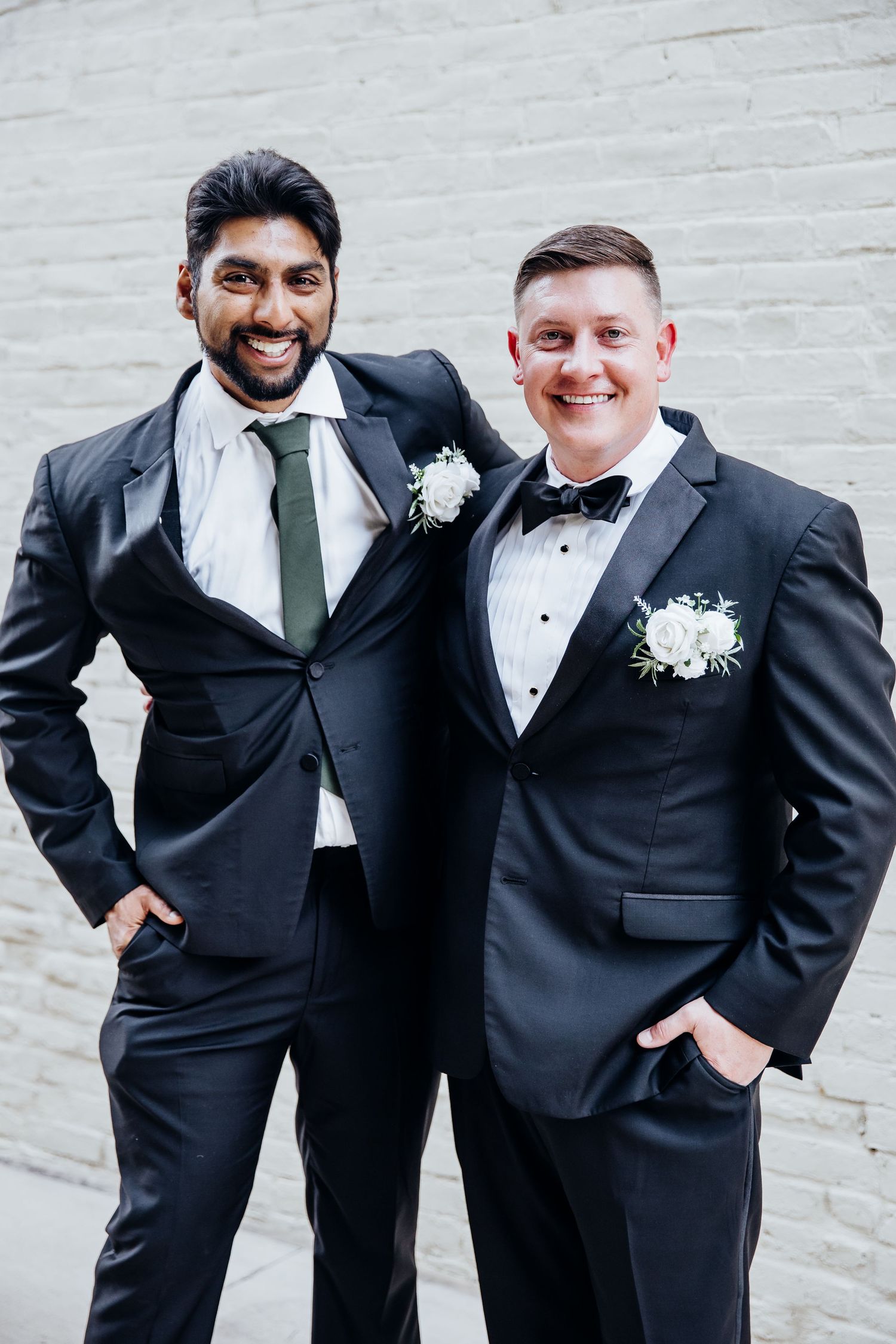 Two people in formal black tuxedos share joyful moments against a white brick wall.