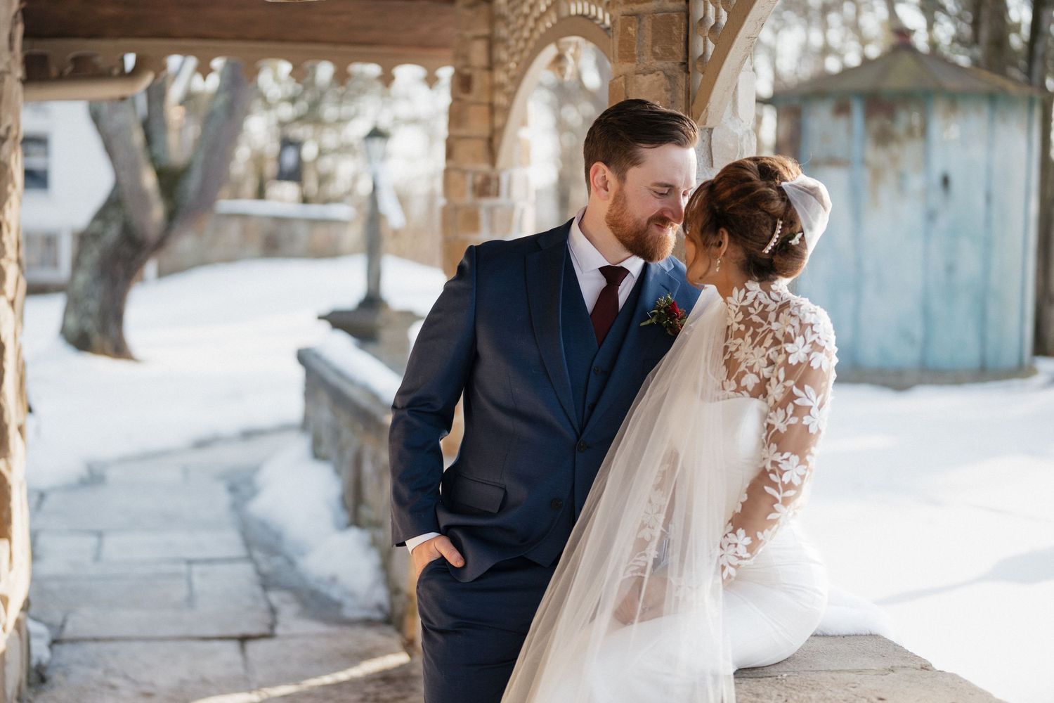 Bride and groom share an intimate moment outdoors in winter, she in a lace gown with veil, he in a navy suit.