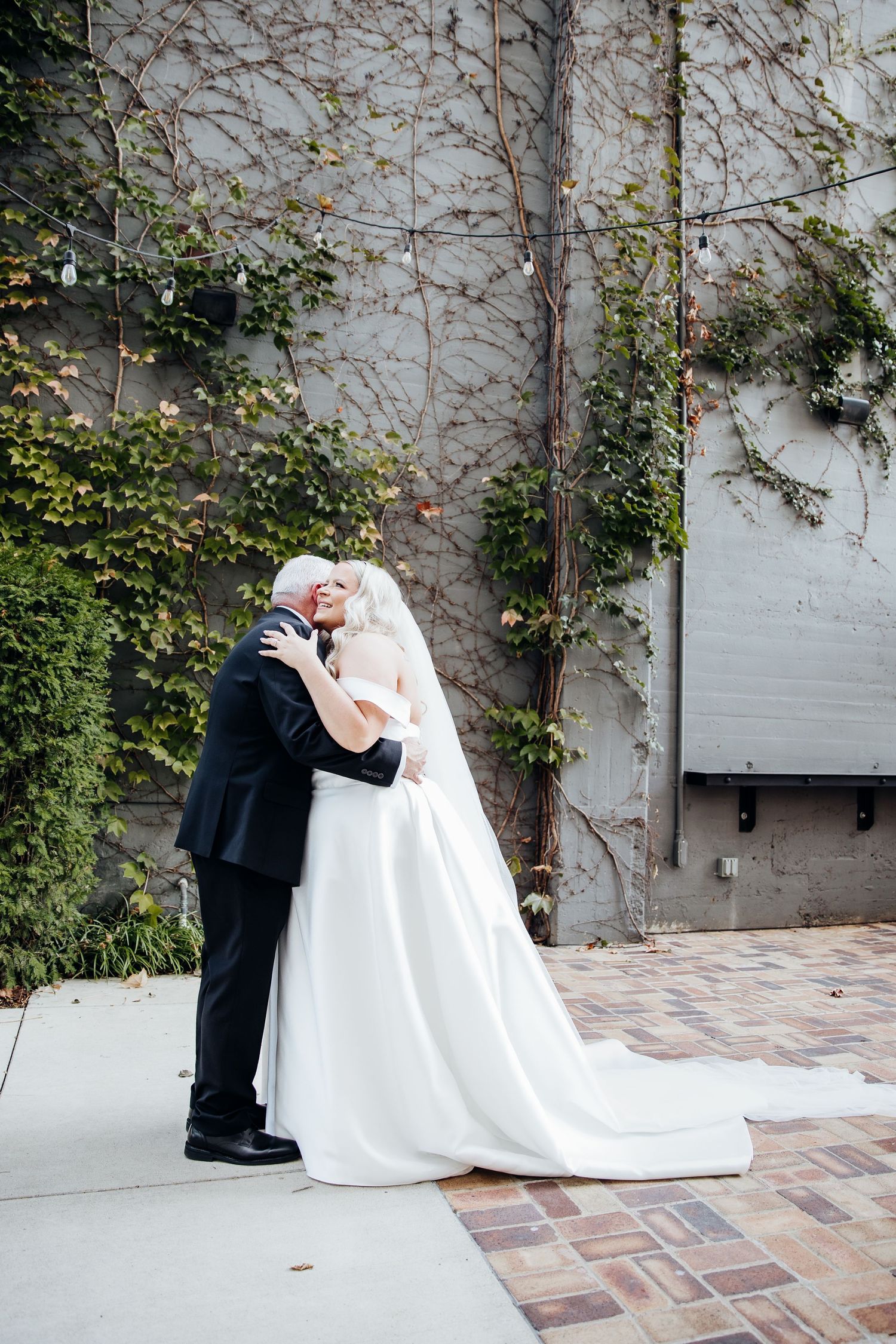 A romantic wedding couple embraces against a vine-covered wall in an outdoor courtyard setting.