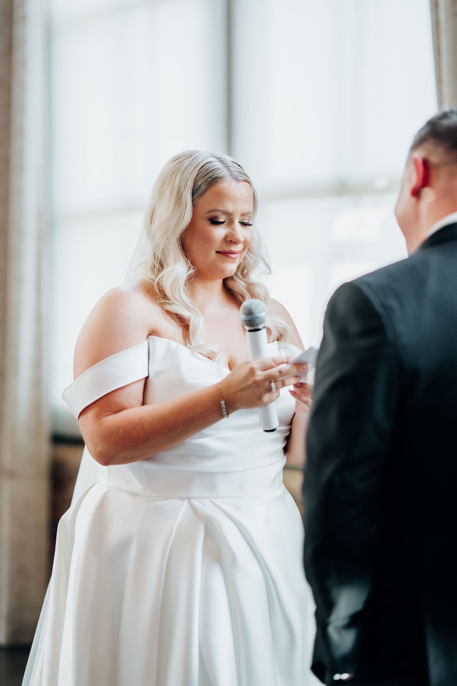 Bride in an elegant off-shoulder white gown holds a microphone during wedding reception speeches.