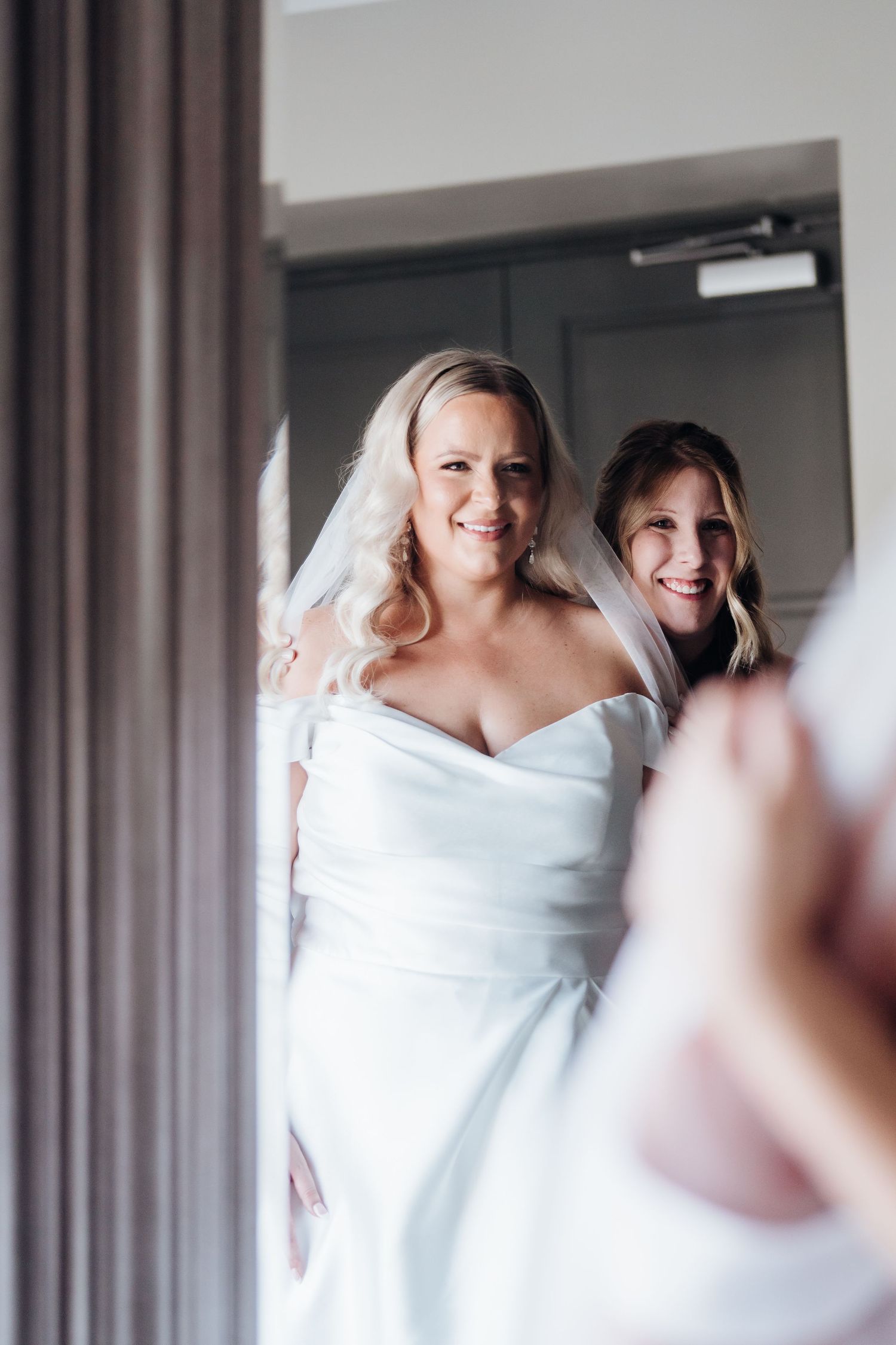 A bride in an off-shoulder white gown smiles while looking in a mirror.