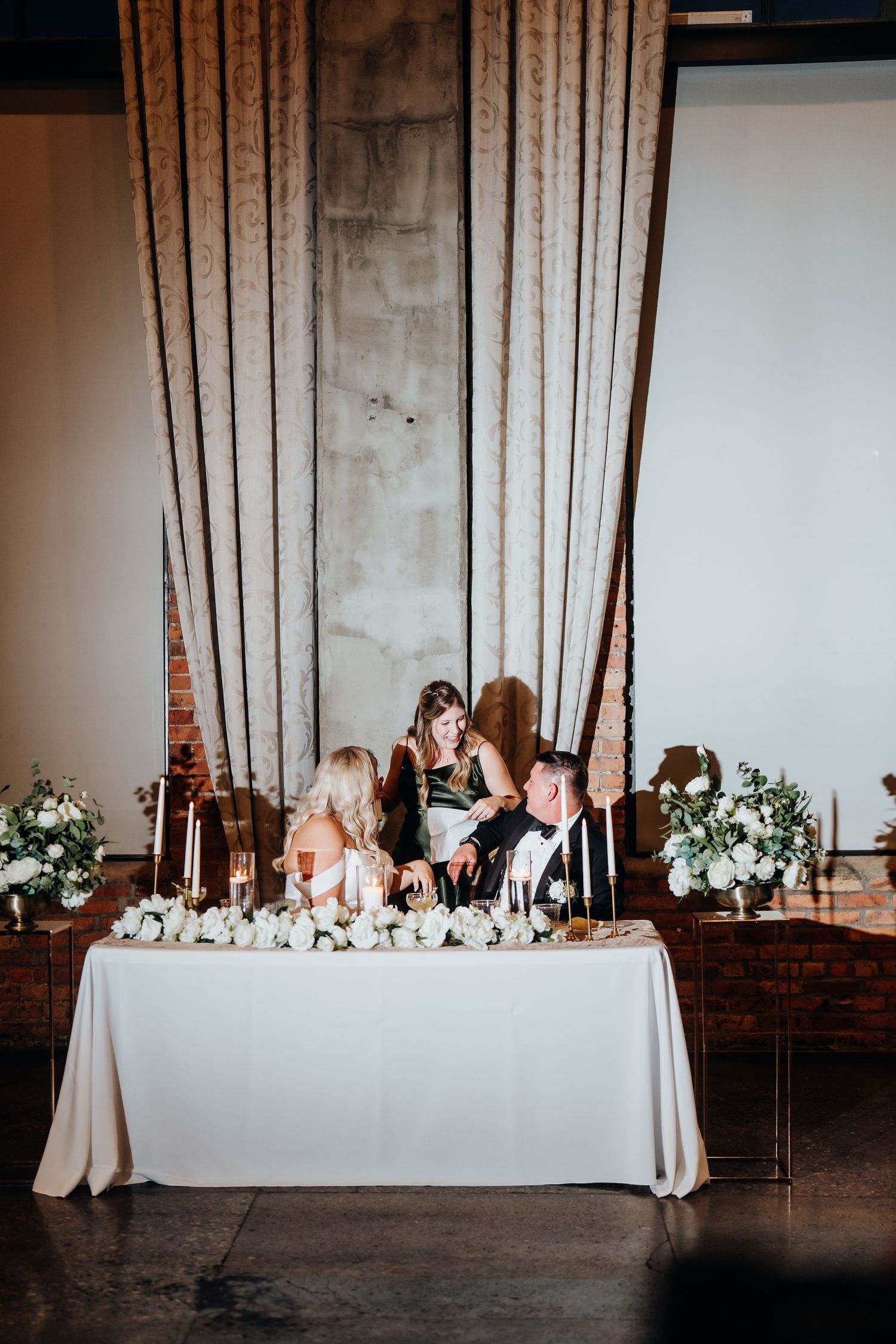 Wedding reception head table with white linens and floral arrangements in industrial venue.