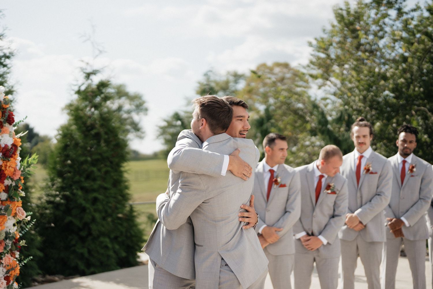 Wedding party members in light gray suits share emotional embrace during outdoor ceremony.