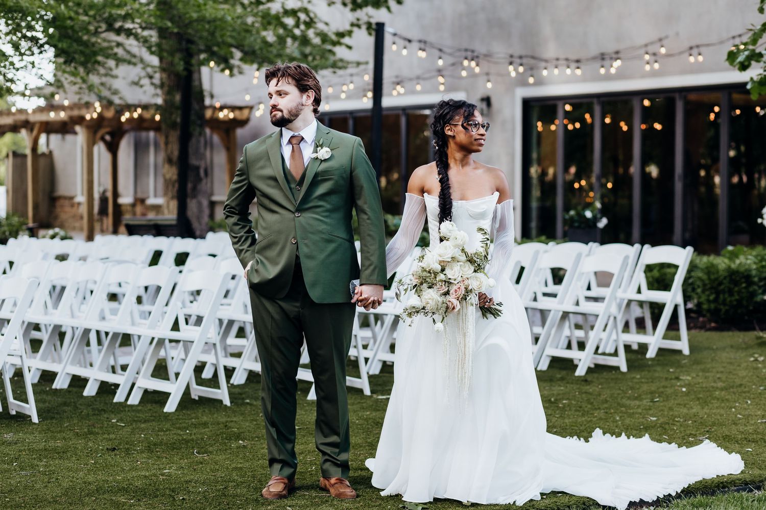 Outdoor wedding ceremony setup with white chairs and string lights in garden setting.
