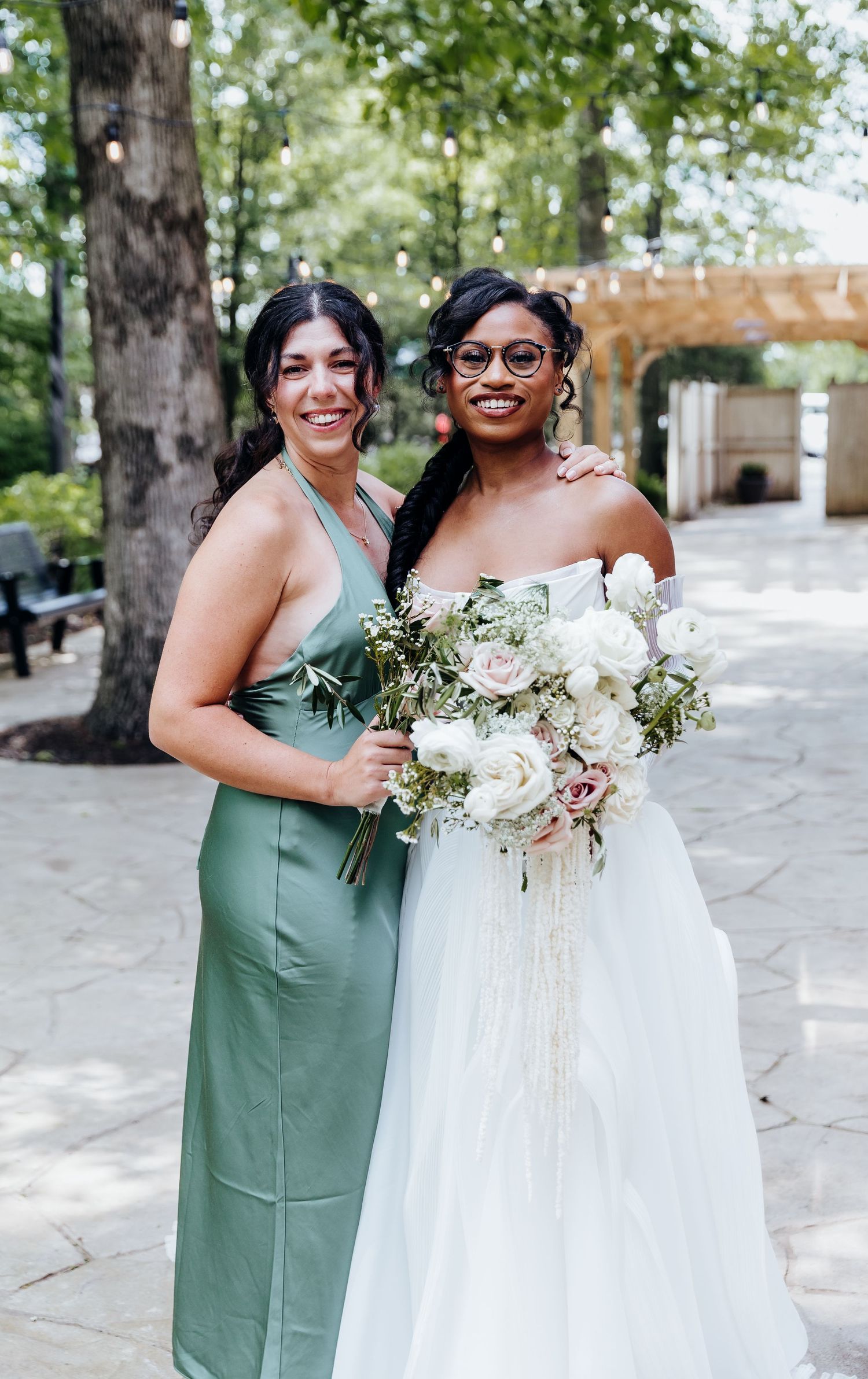 Wedding party members in sage green dresses and white gown pose with elegant floral bouquets outdoors.