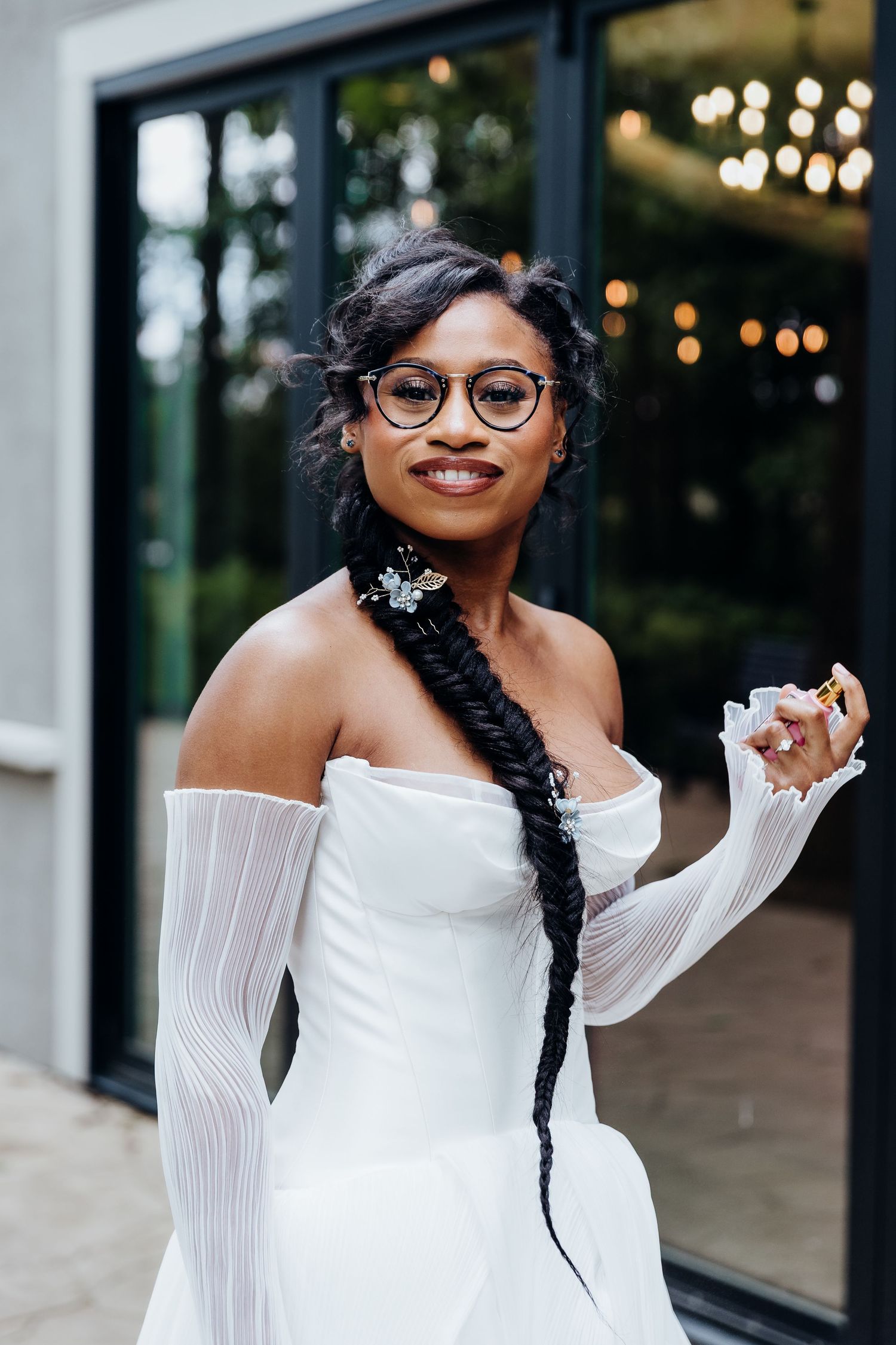 A beautiful wedding look featuring an off-shoulder white dress with flowing sleeves, styled with modern glasses and a long braid.