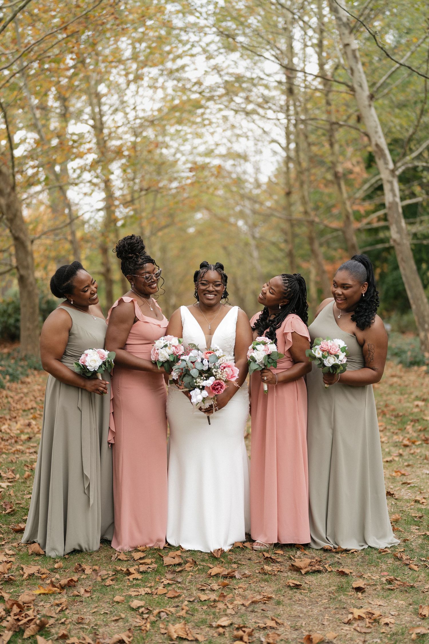 Bridal party in alternating pink and gray dresses pose together on autumn leaves.