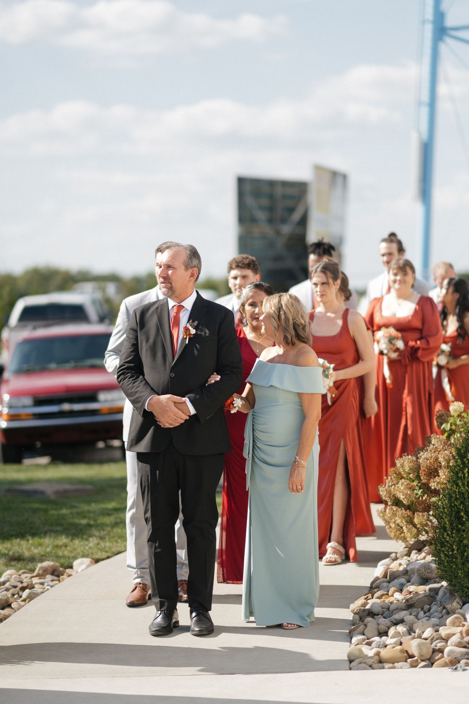 Wedding processional with attendants in red dresses walking down outdoor path.