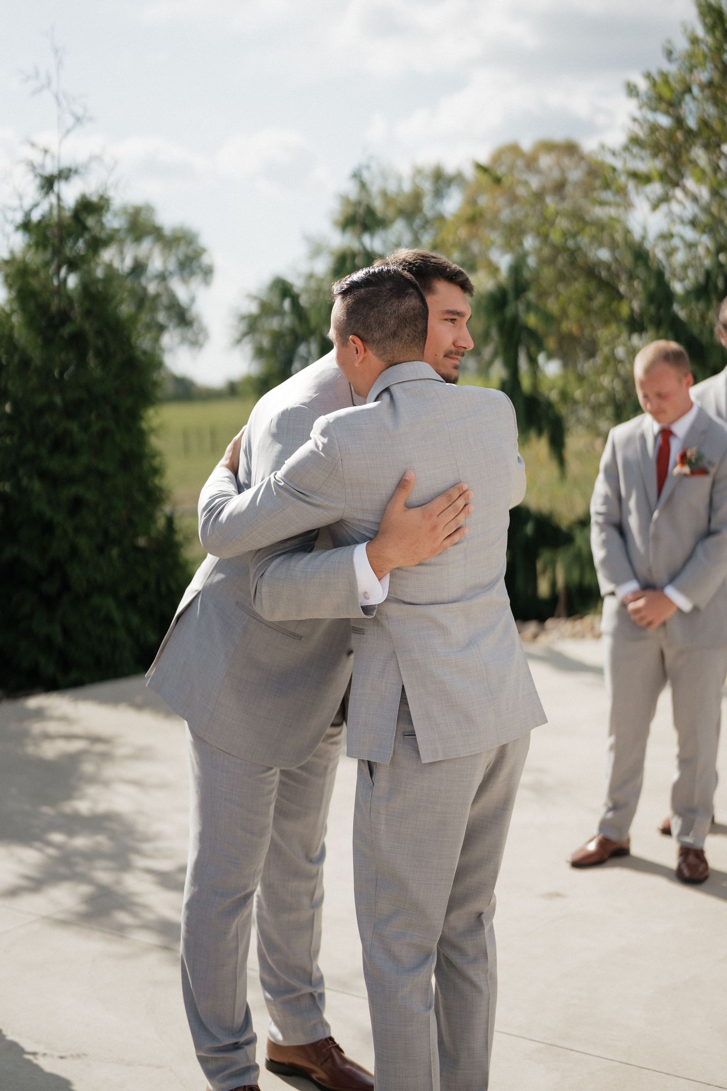 Two people in light gray suits share an emotional embrace during an outdoor wedding ceremony.