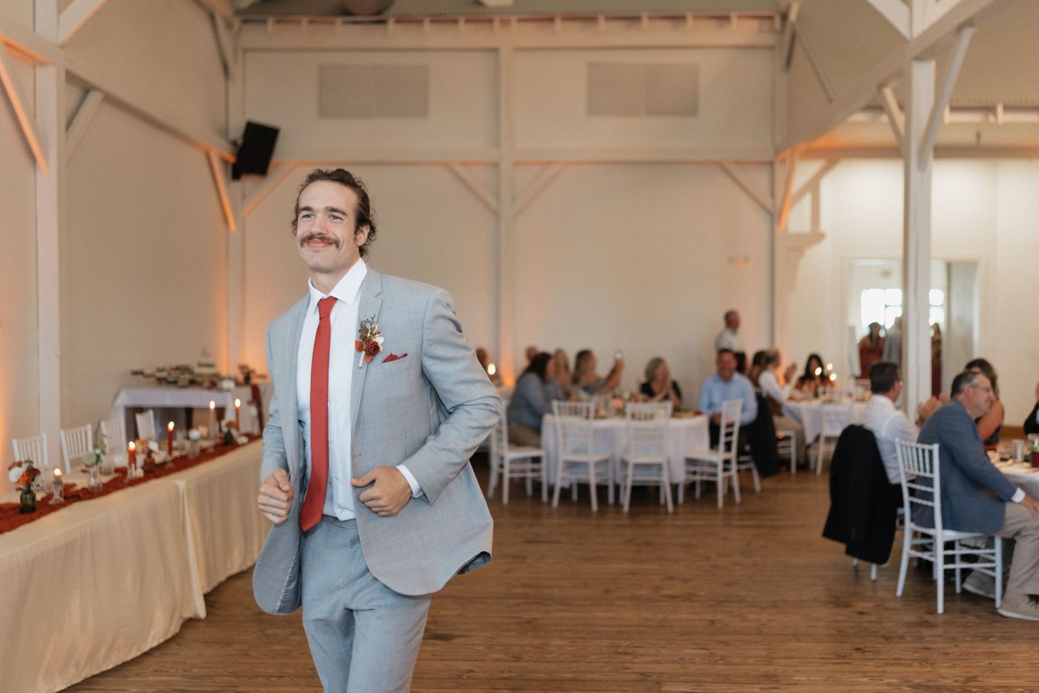 Wedding guest dances enthusiastically in gray suit at reception venue with exposed beams.