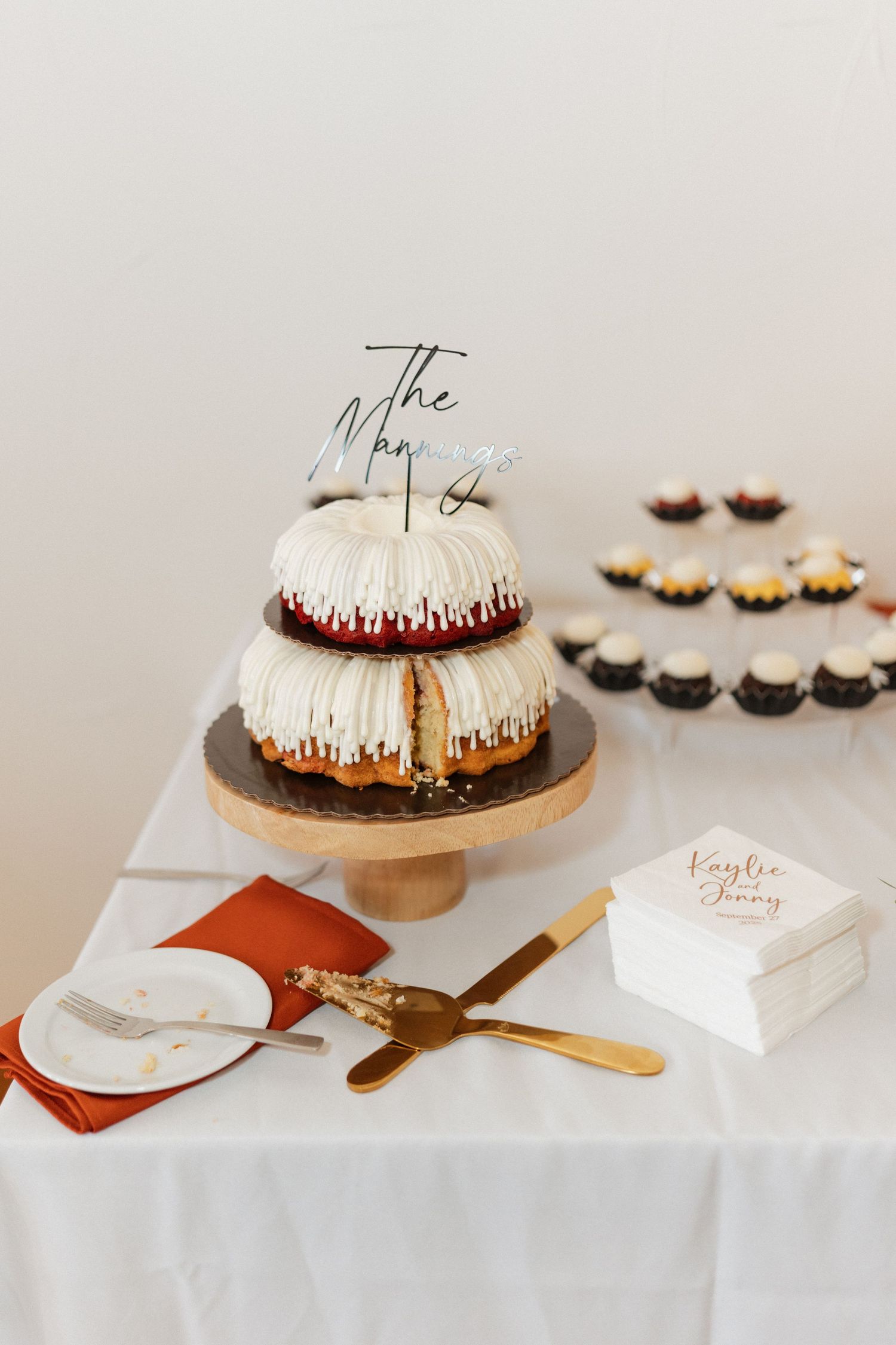 Modern tiered wedding cake with cake topper displayed on wooden stand with serving utensils.