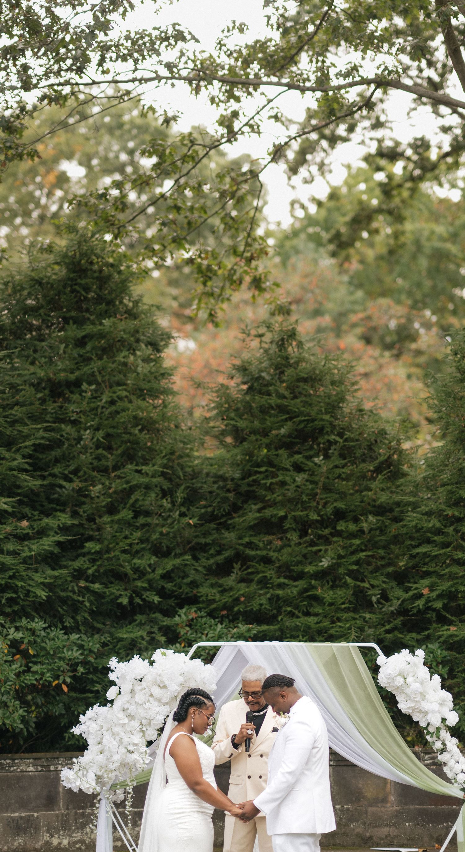 Wedding ceremony under white floral arch with evergreen trees in background.