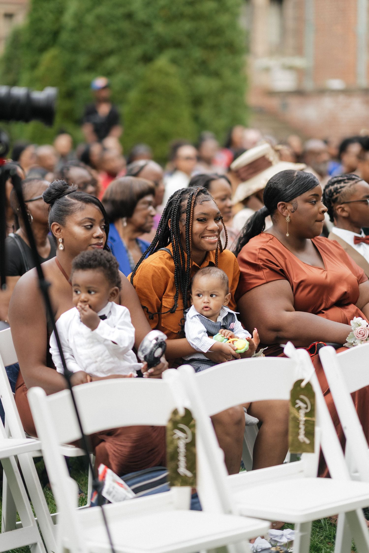 Wedding guests seated in white chairs smile and watch the ceremony outdoors.