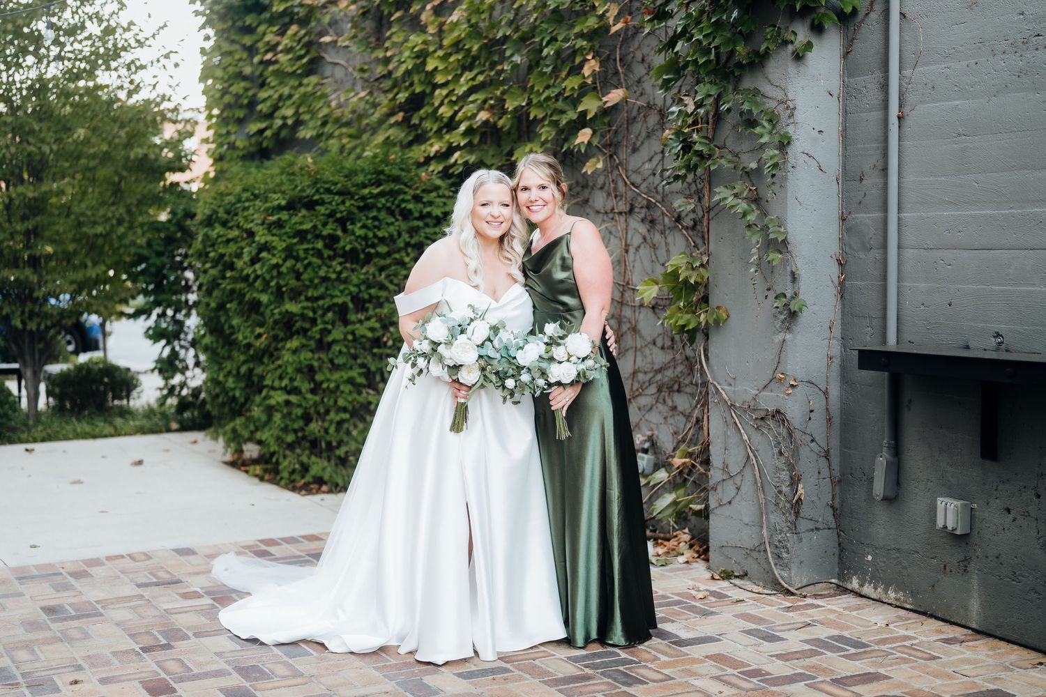 Two people in wedding attire pose together against an ivy-covered wall in an outdoor garden setting.