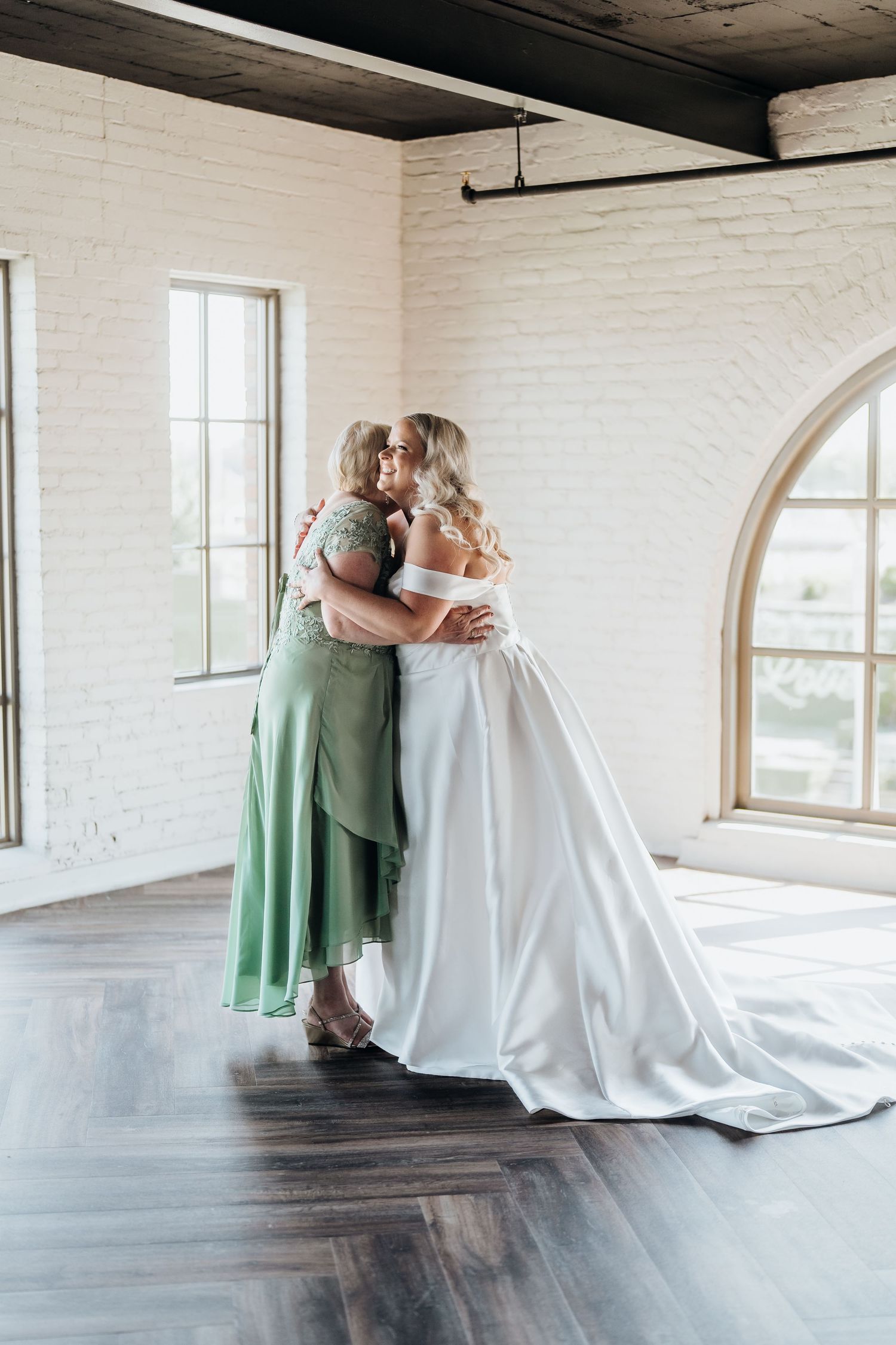 Emotional first look moment between bride in white gown and bridesmaid in sage green dress in bright loft space.