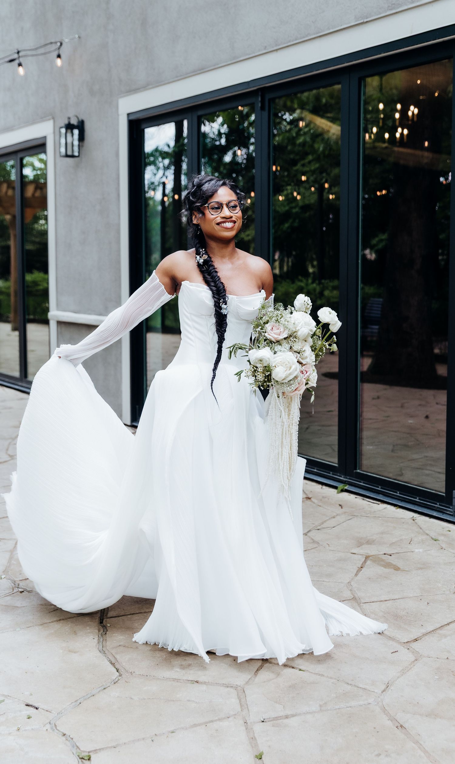 A bride in a flowing white gown holds her dress while carrying a white floral bouquet outdoors.