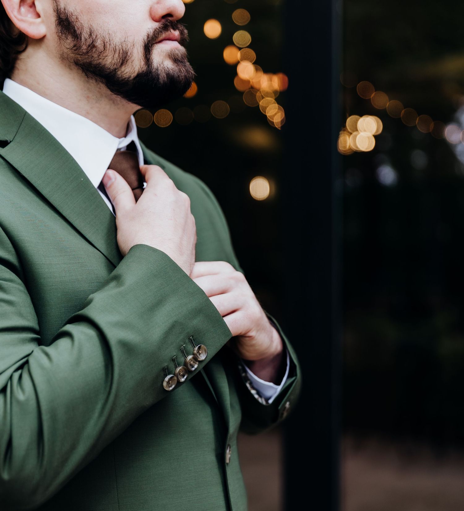 Man wearing olive green suit jacket adjusting collar against dark background with bokeh lights.
