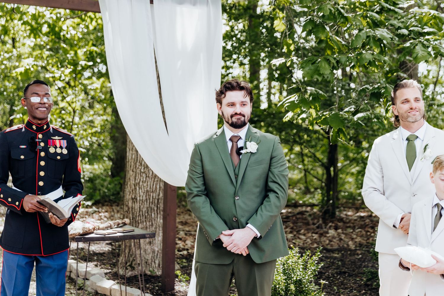 Wedding party poses for formal photos in an outdoor garden setting with white draping and greenery.