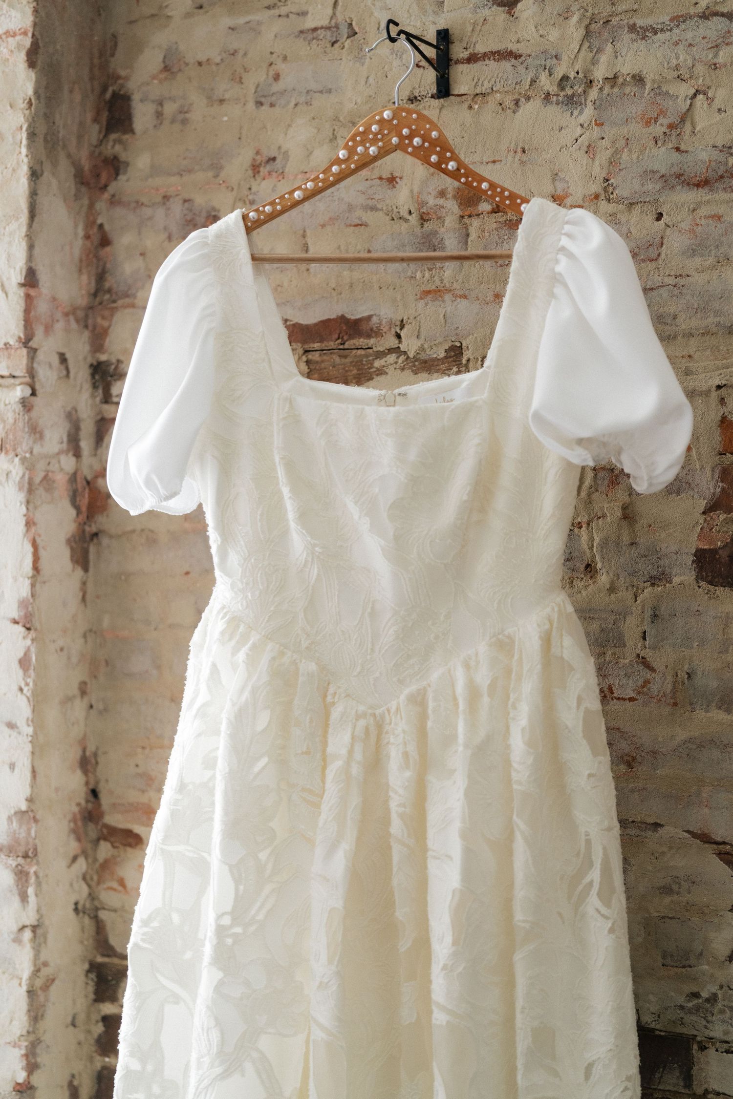 Ivory wedding dress with puff sleeves and lace skirt on pearl-adorned wooden hanger against rustic brick wall.