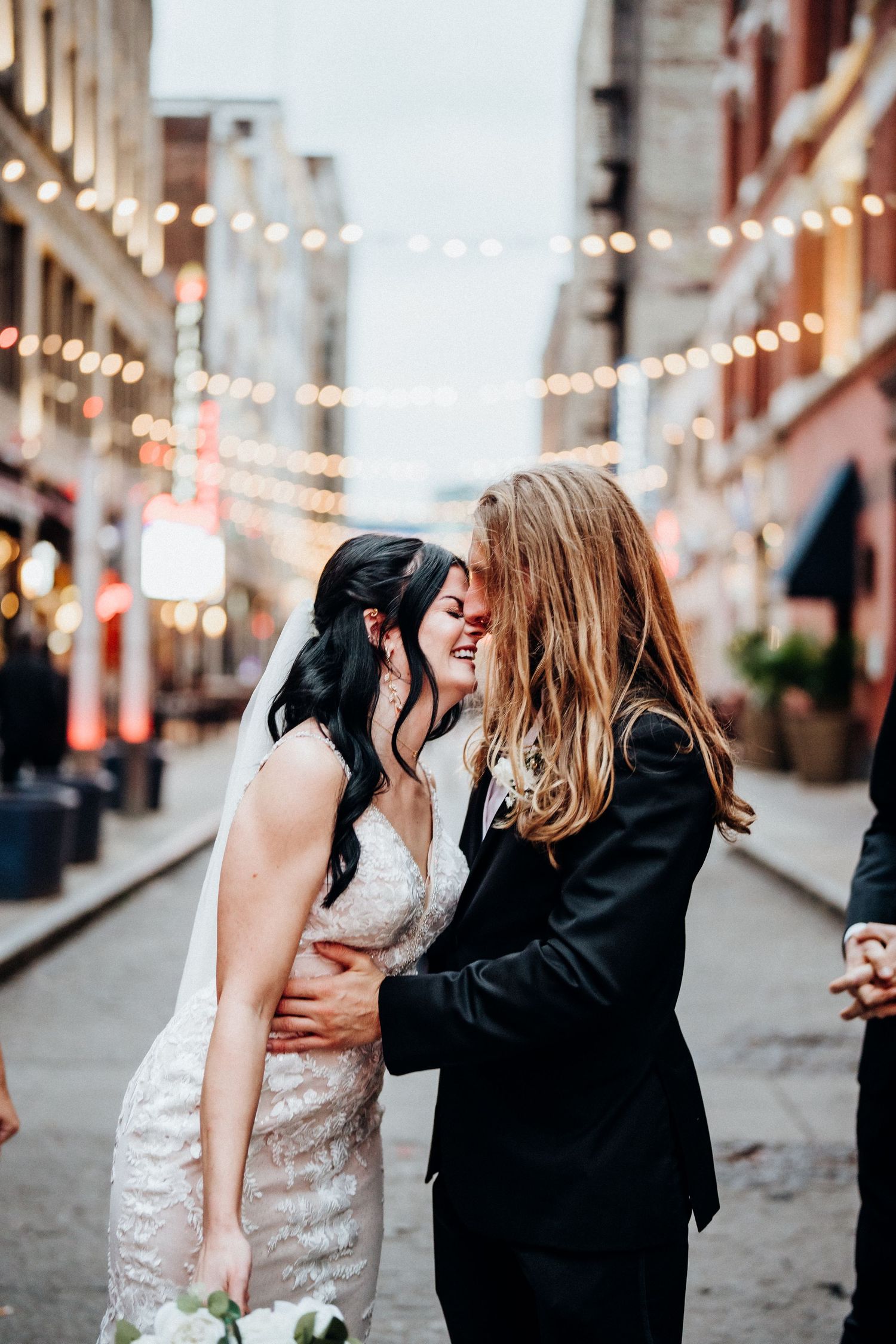 A romantic moment under string lights in an urban setting with brick buildings.