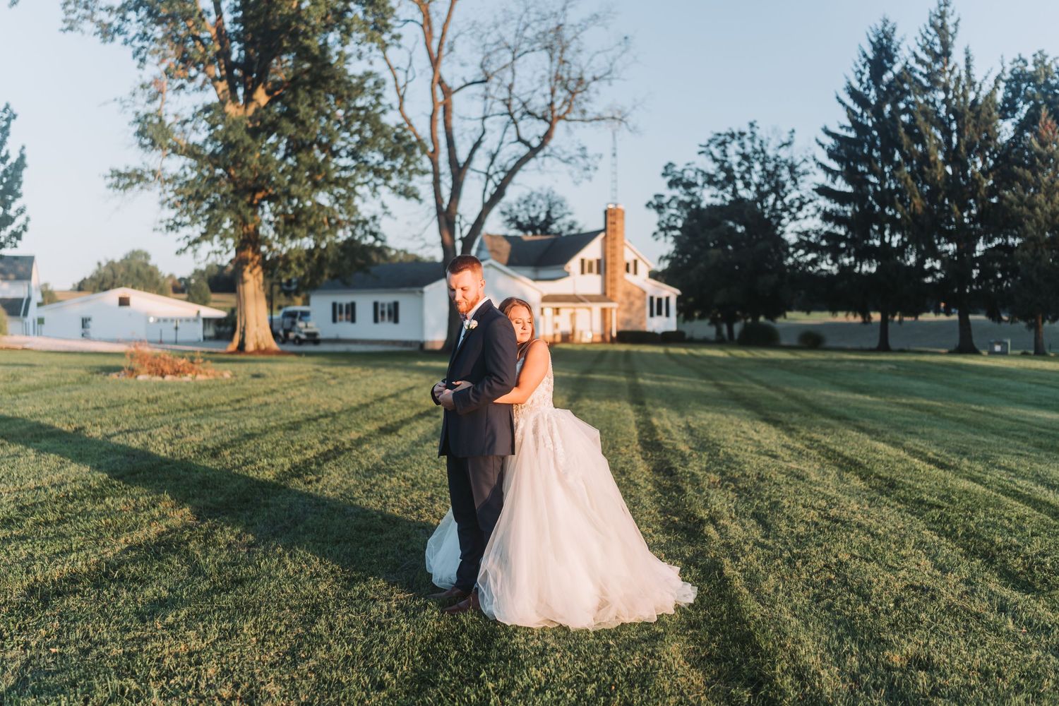 Wedding couple embracing in golden sunlight on sprawling lawn with buildings in background.