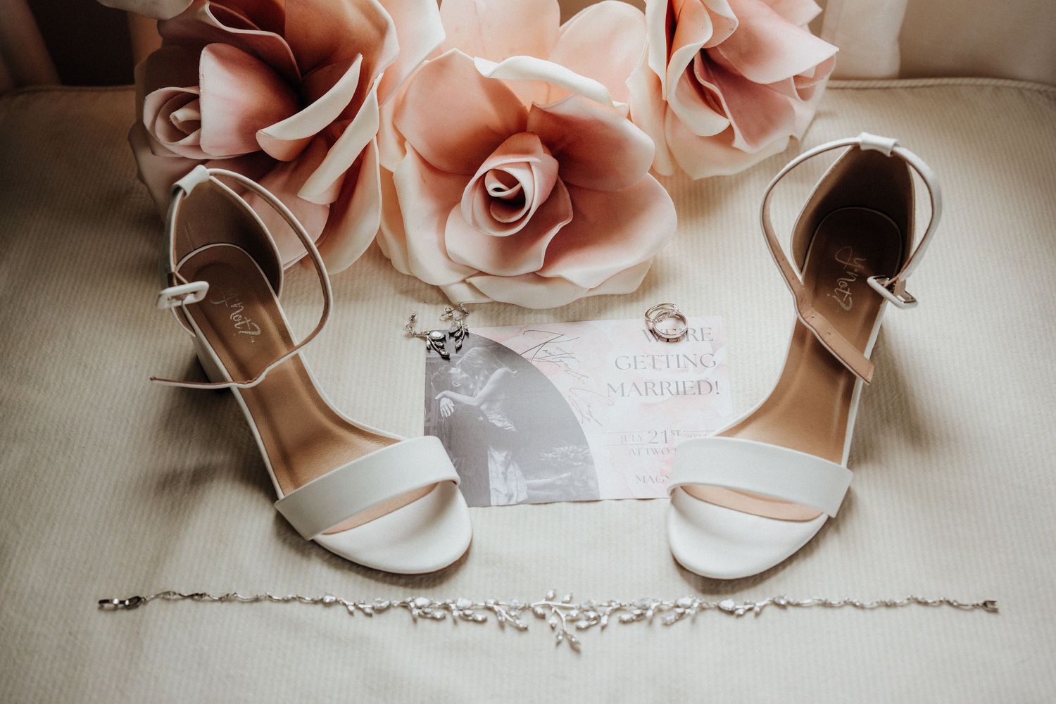 Elegant white wedding shoes and delicate pink roses displayed with wedding rings and invitation.