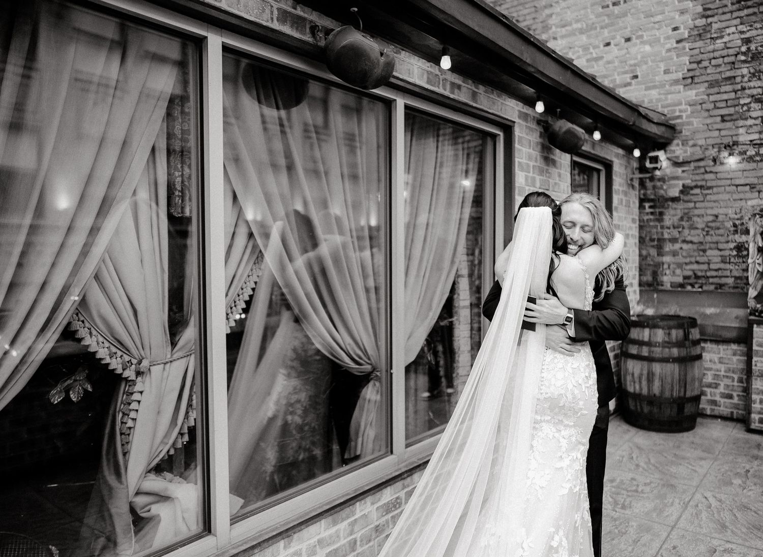 A romantic black and white photo of a couple embracing near sheer curtained windows.