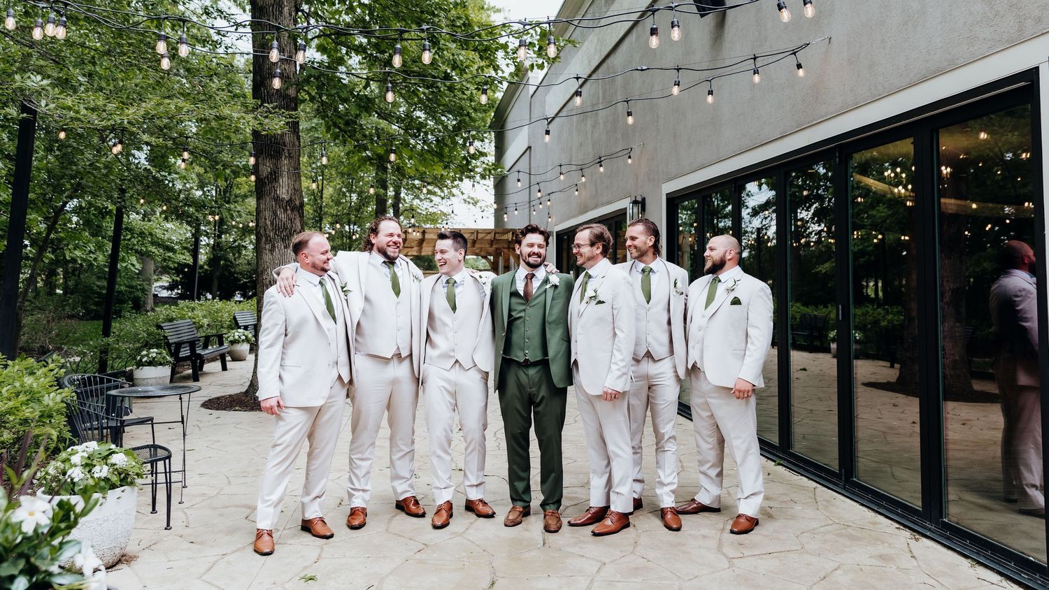 Wedding party members in white and green suits stand in line against a modern building exterior.